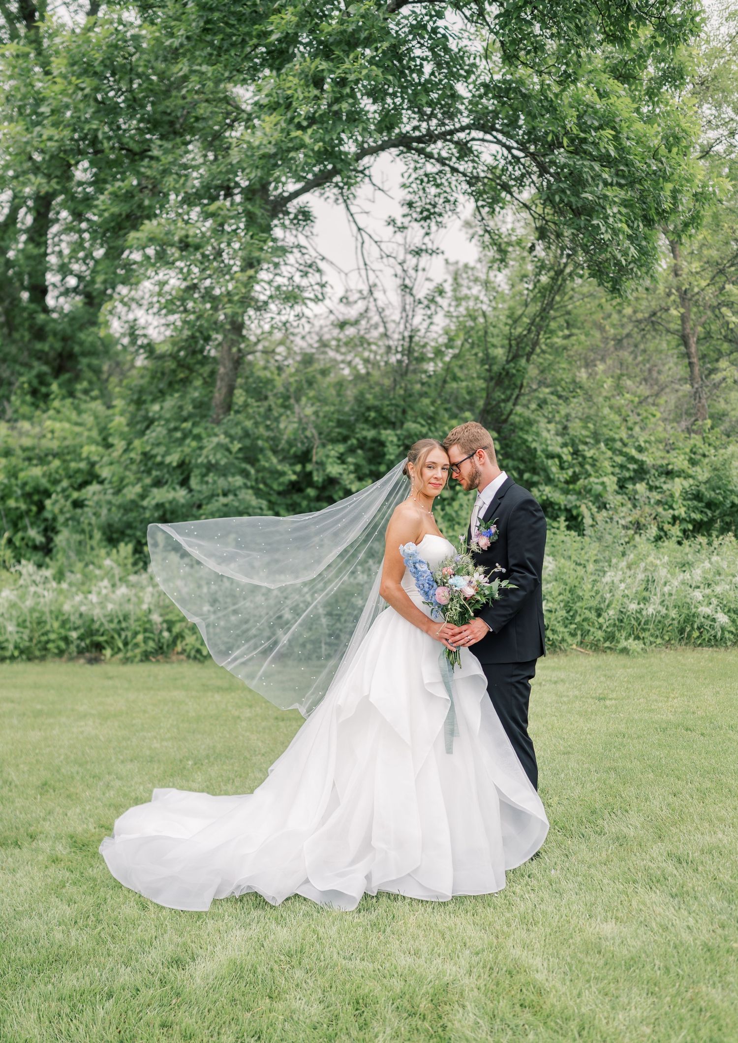 A romantic outdoor wedding photo with a flowing veil and white gown against lush green foliage in a garden setting.