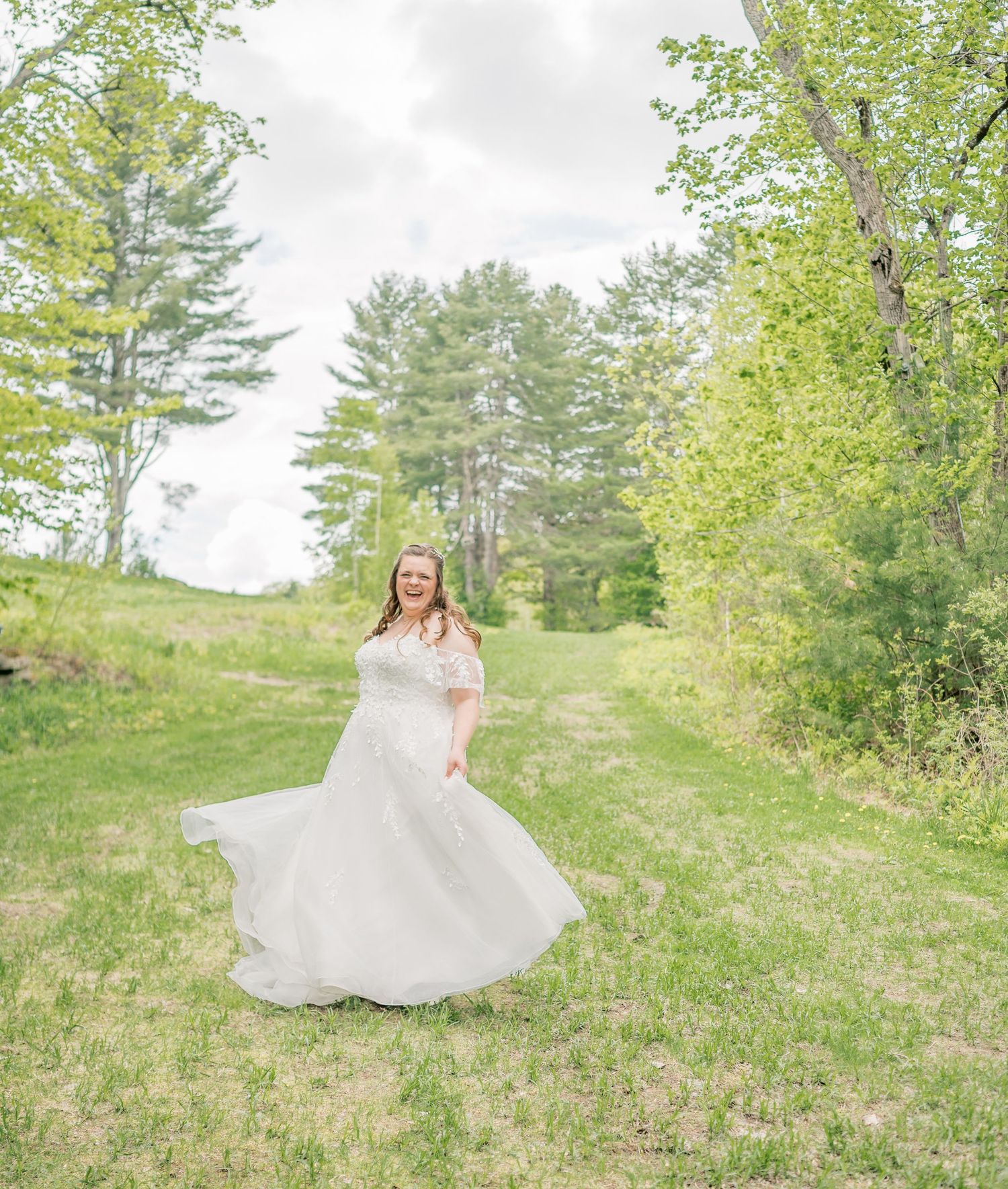 A flowing white wedding dress twirls in a dreamy outdoor setting with evergreen trees and spring foliage in the background.