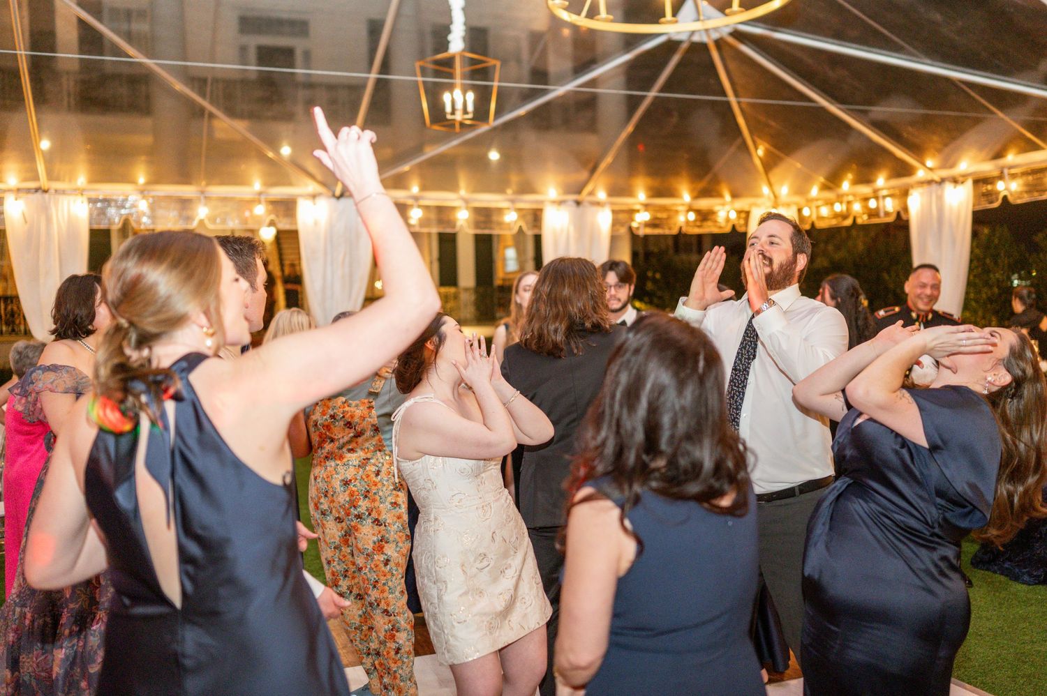 Wedding guests dance and celebrate with their hands in the air under twinkling lights at an evening reception.