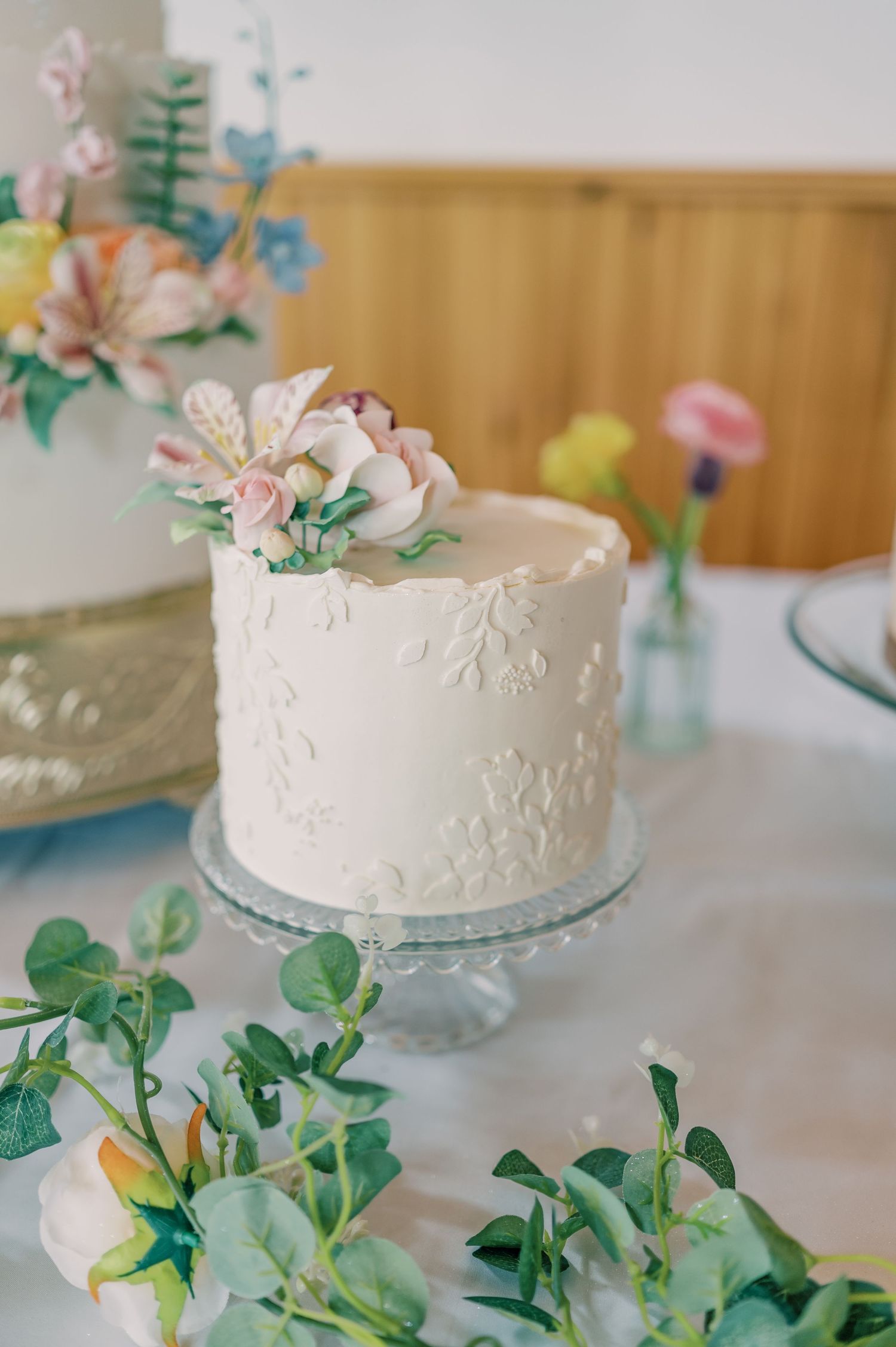 White frosted wedding cake with delicate floral details displayed on glass pedestal with greenery decorations.