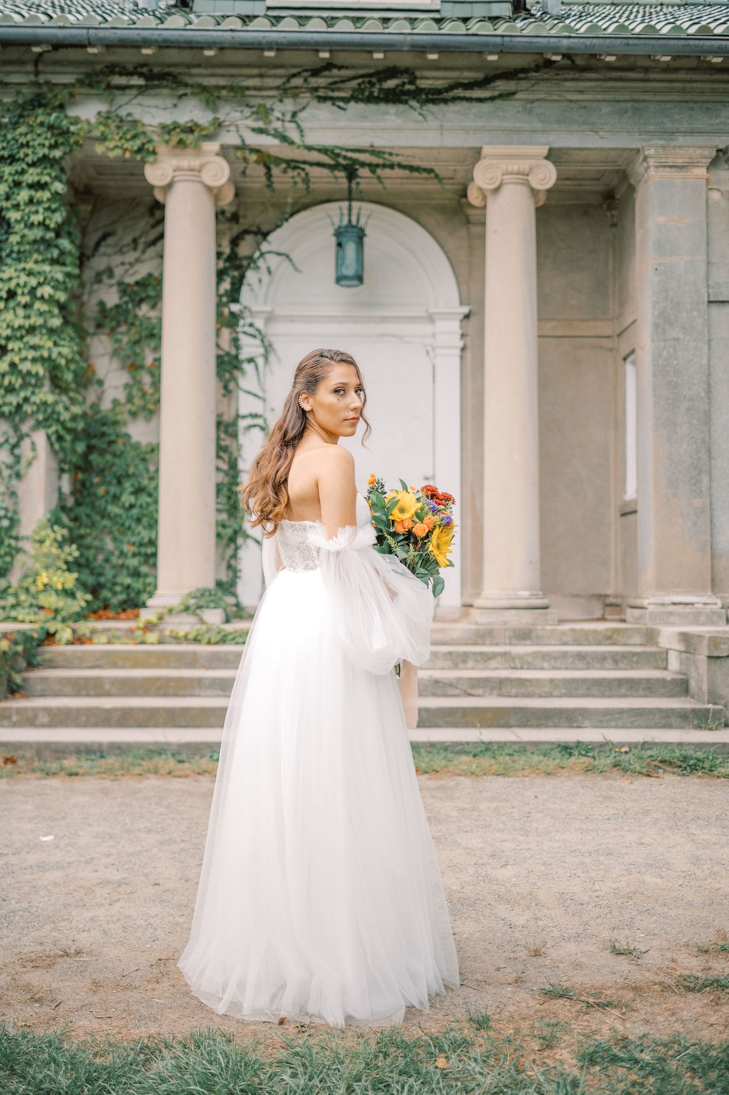 Elegant bride in flowing white tulle gown poses gracefully in front of classical stone columns at outdoor venue.