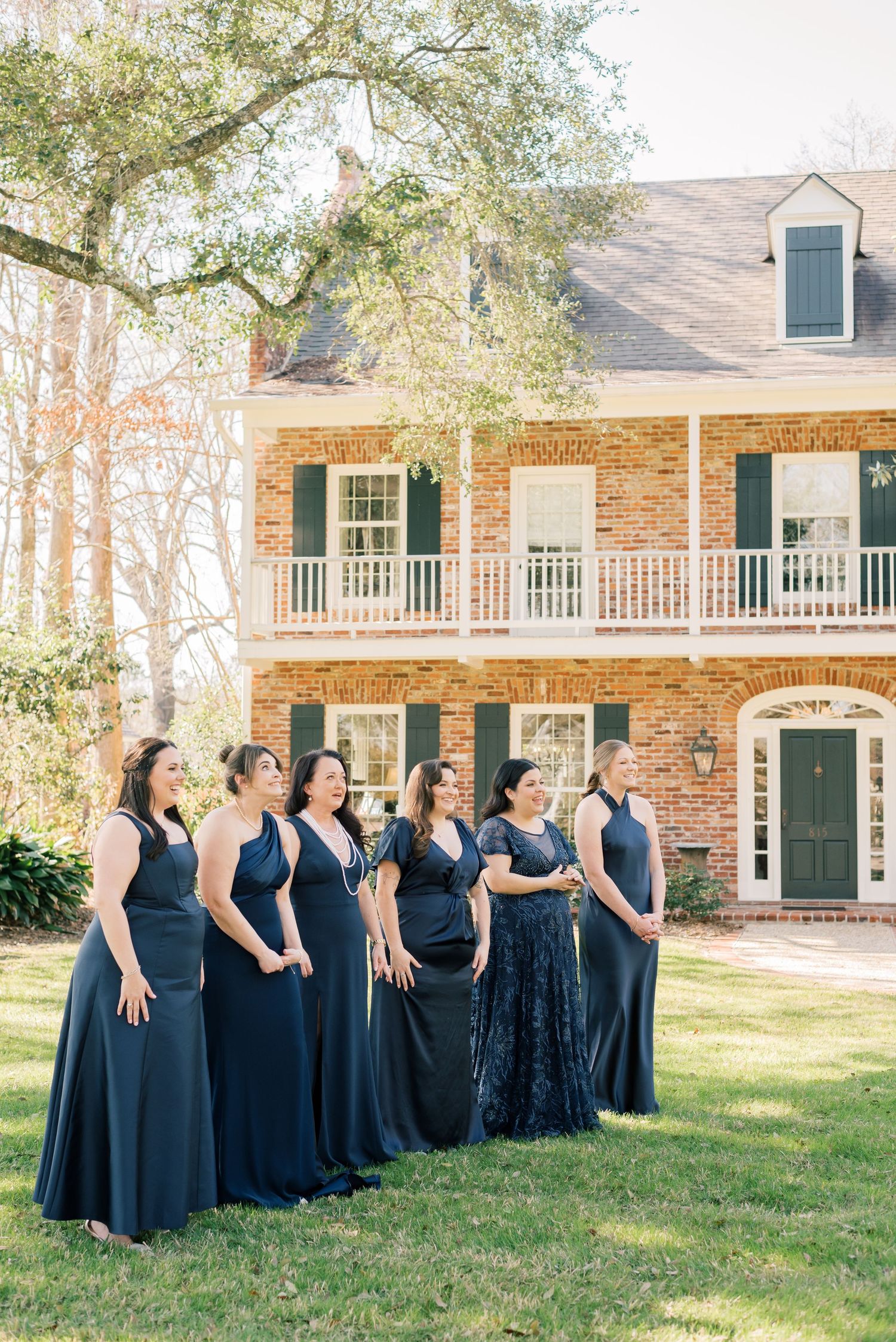 A group of people wearing navy blue dresses stand in a line on a lawn in front of a brick colonial-style house.