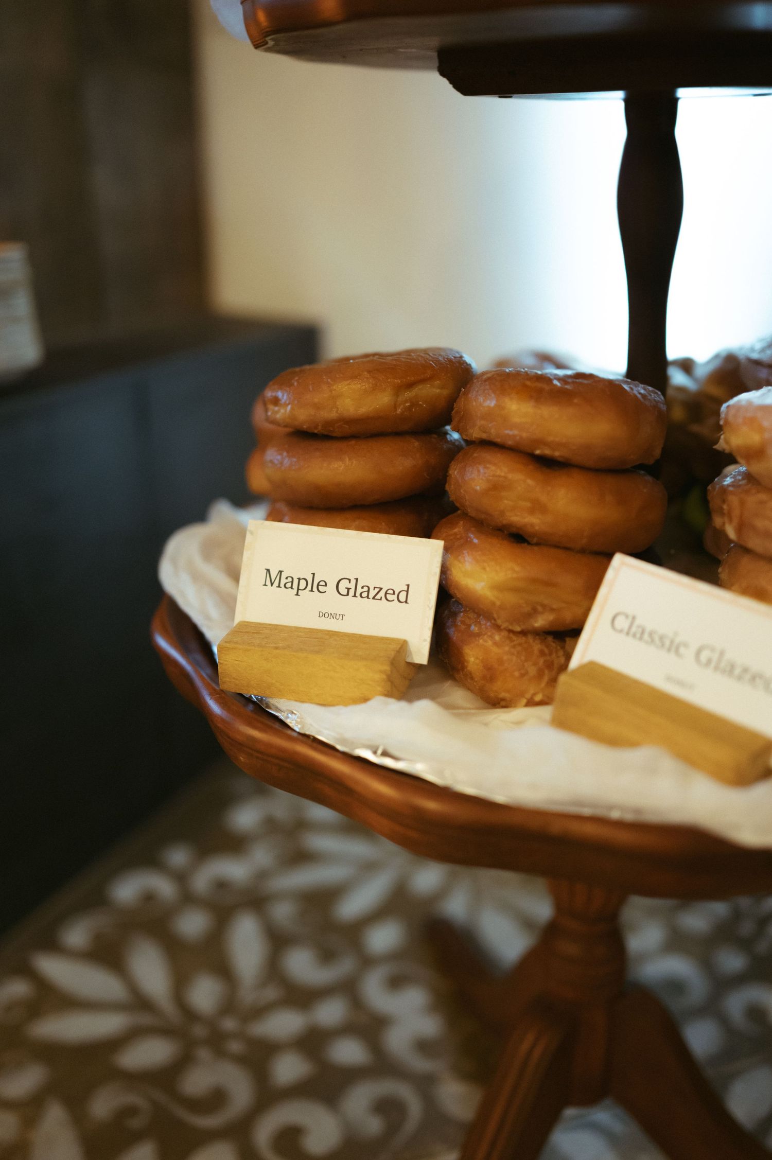 Maple glazed donuts displayed on a wooden tiered serving stand with small identifying labels.
