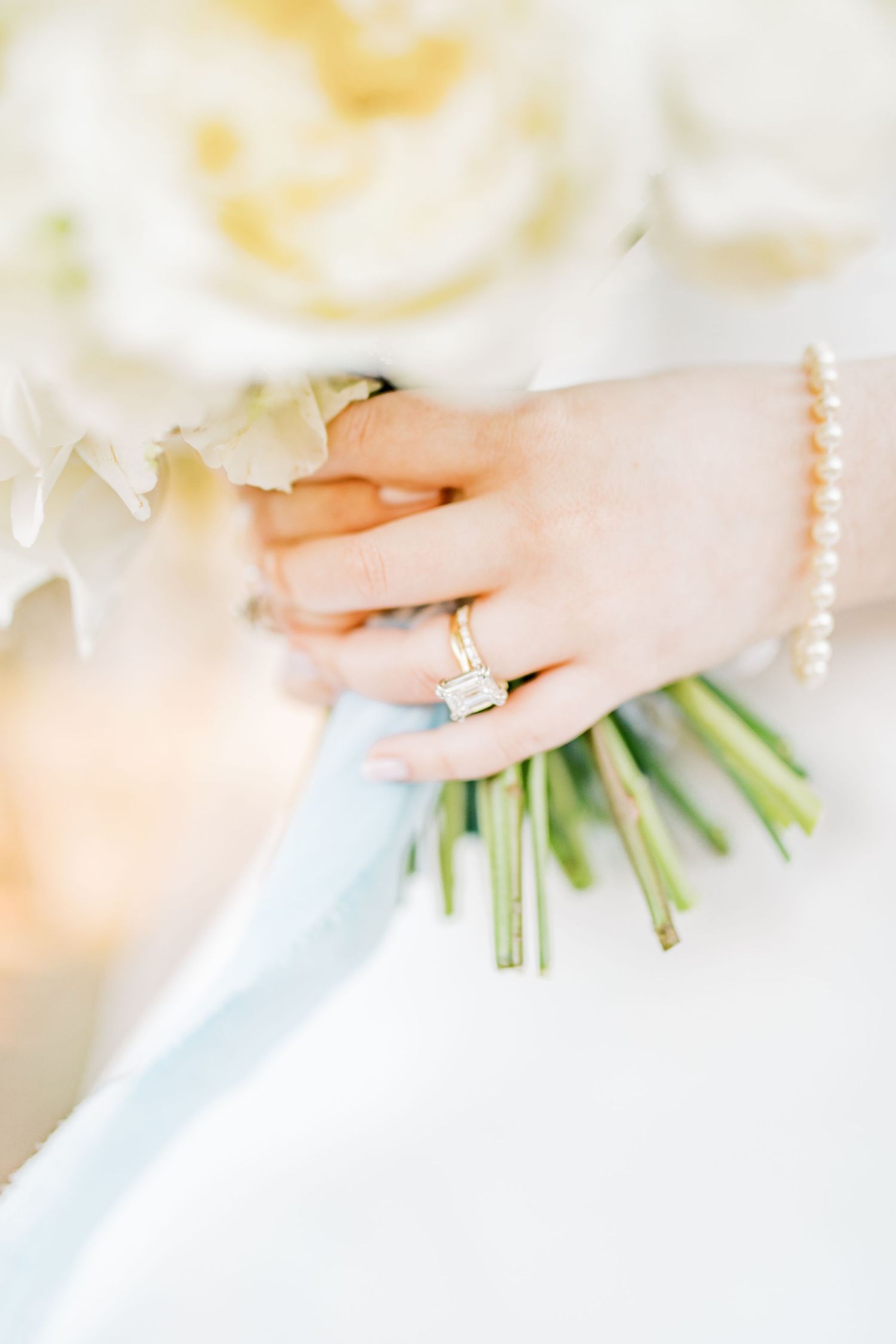 Close-up of hands holding white bouquet with blue ribbon and engagement ring detail in soft natural light.