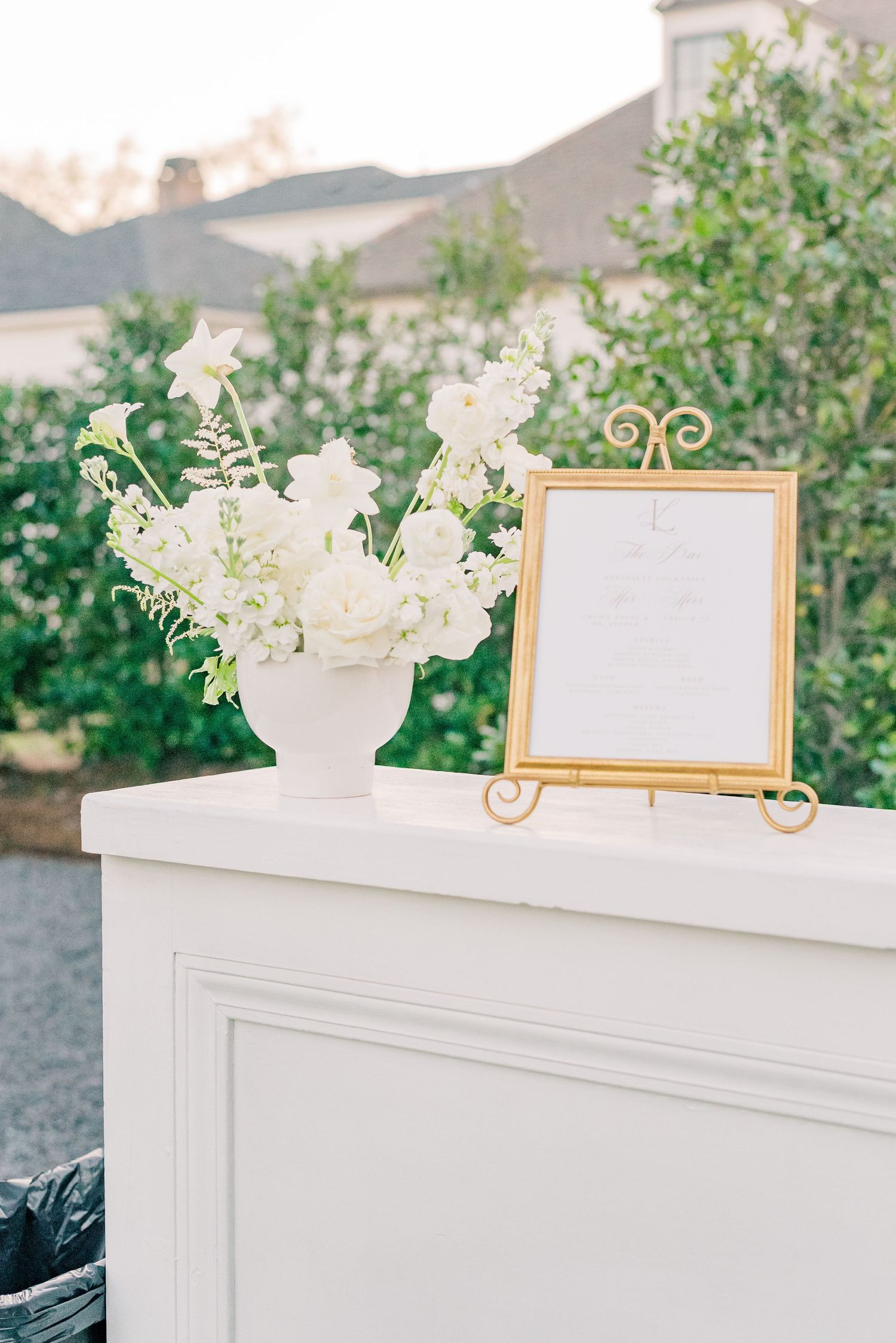 Elegant white floral arrangement in a classic urn vase next to gold-framed sign on white pedestal display.