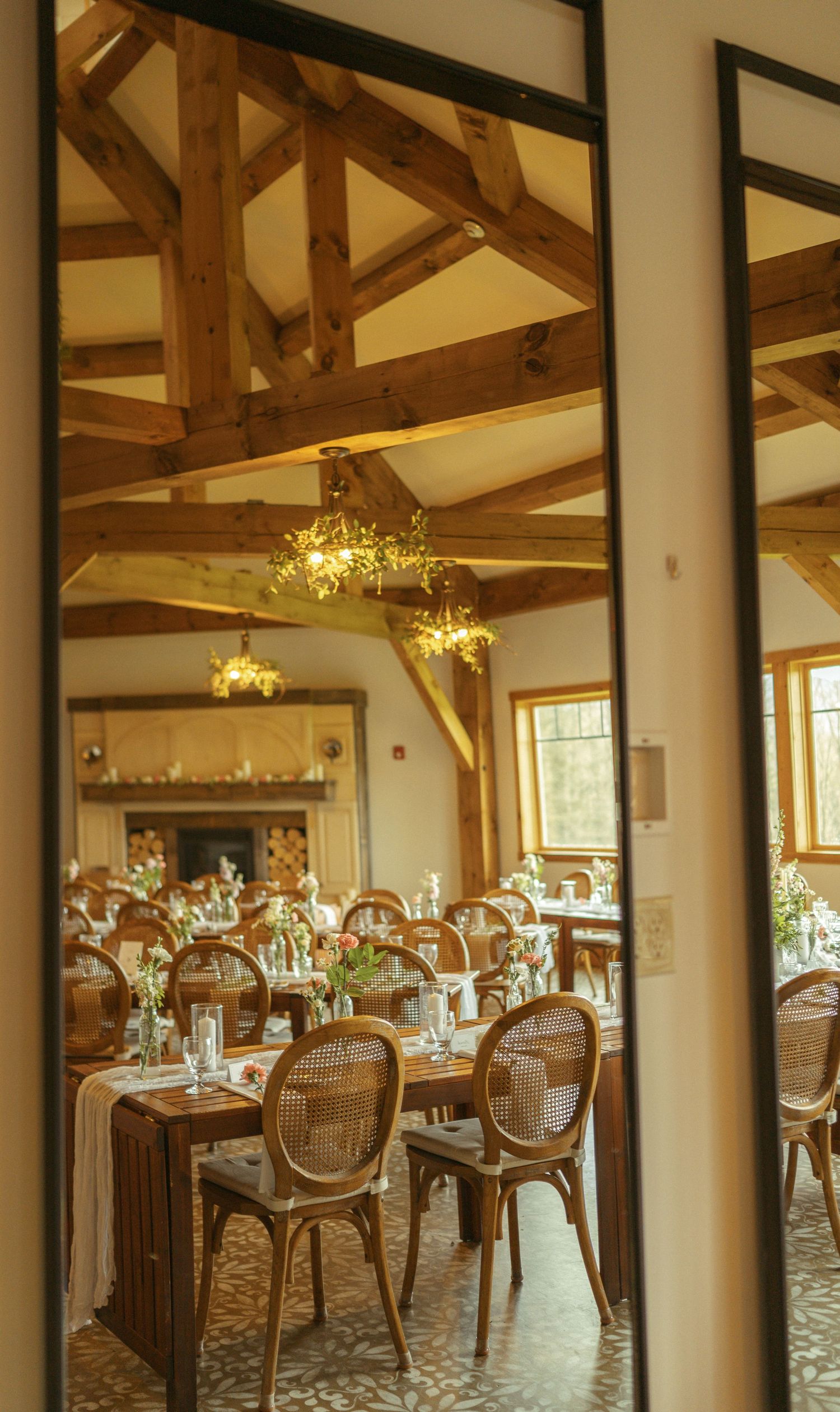 Elegant wedding reception venue with wooden beams, chandeliers and tables set with formal place settings in rustic style.