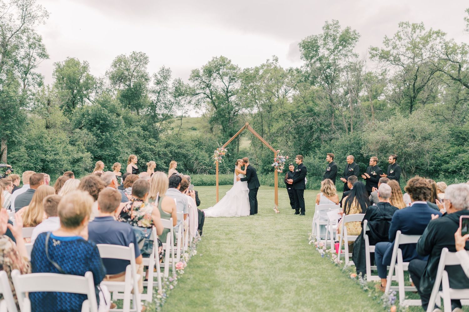 A panoramic view of an outdoor wedding ceremony with guests seated on white chairs in a lush green garden setting.