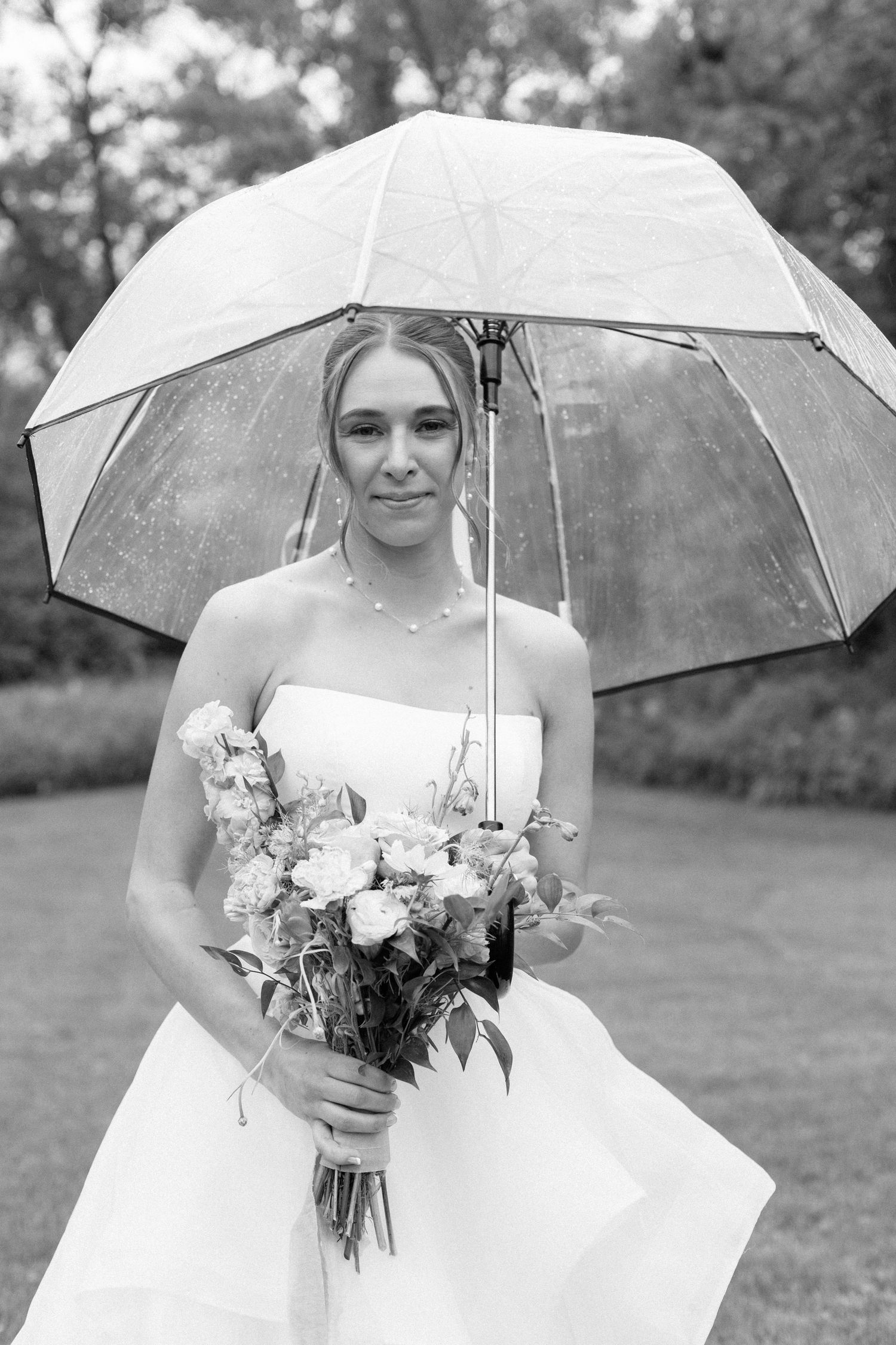 Bride in white wedding dress holds floral bouquet under clear umbrella during outdoor photo session in garden setting.