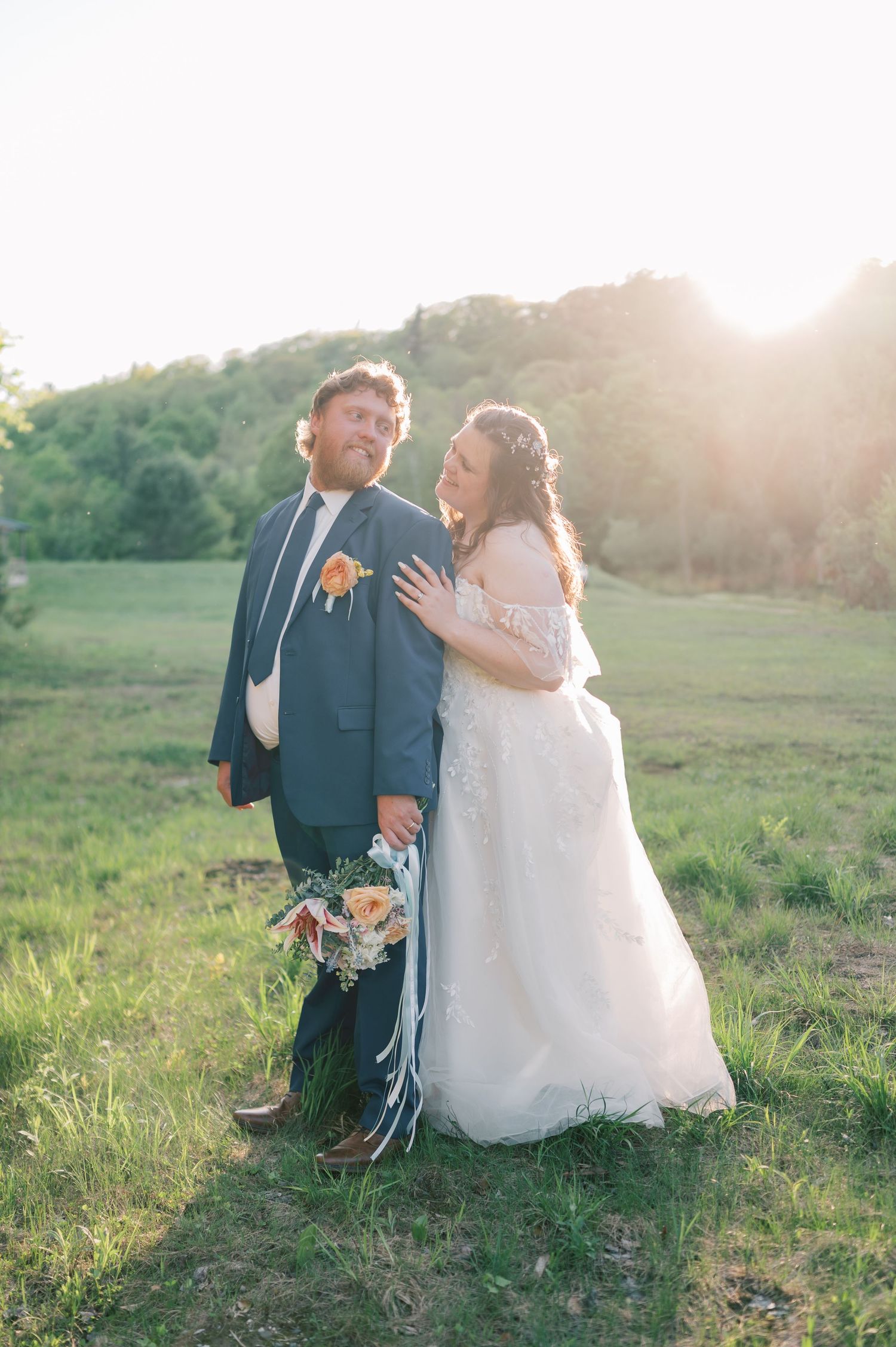 A couple in wedding attire sits together in a sunlit meadow during golden hour.