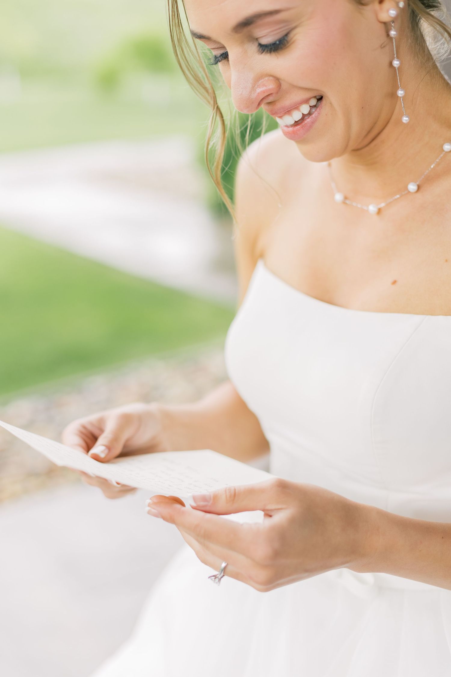 A bride in a white off-shoulder dress reads a letter while smiling on her wedding day.