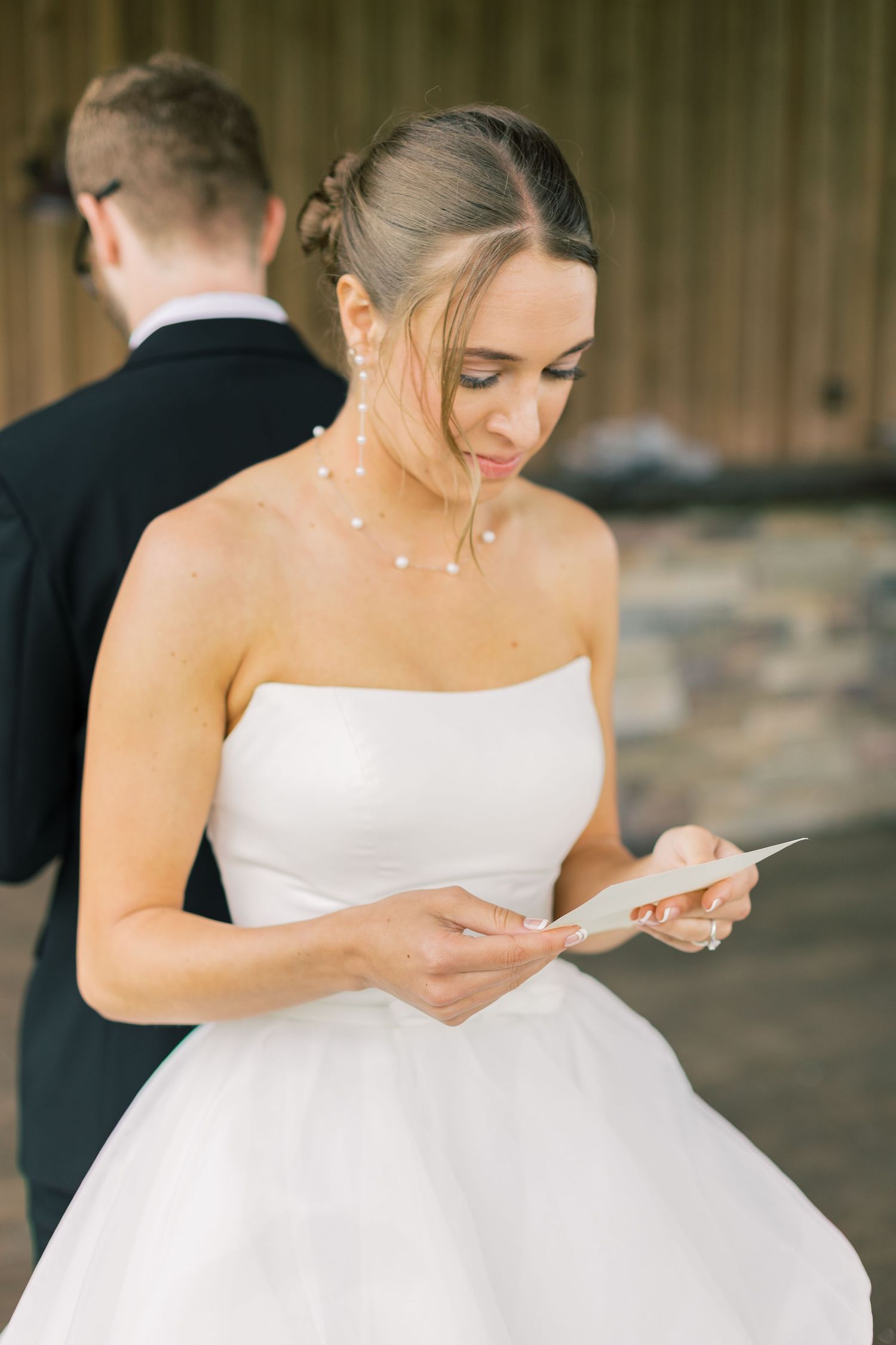 Bride in strapless white ballgown reads a note while wearing delicate pearl hair accessories at outdoor wedding venue.