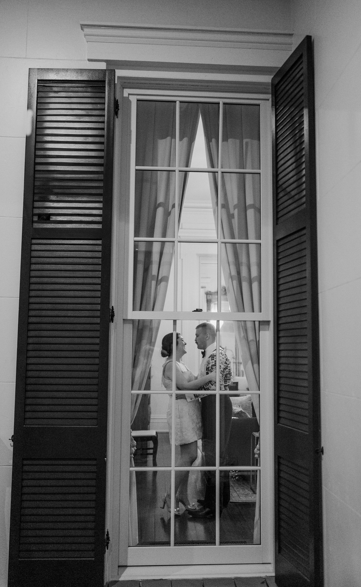 Black and white architectural detail of traditional windows with shutters and white curtains on a building exterior.