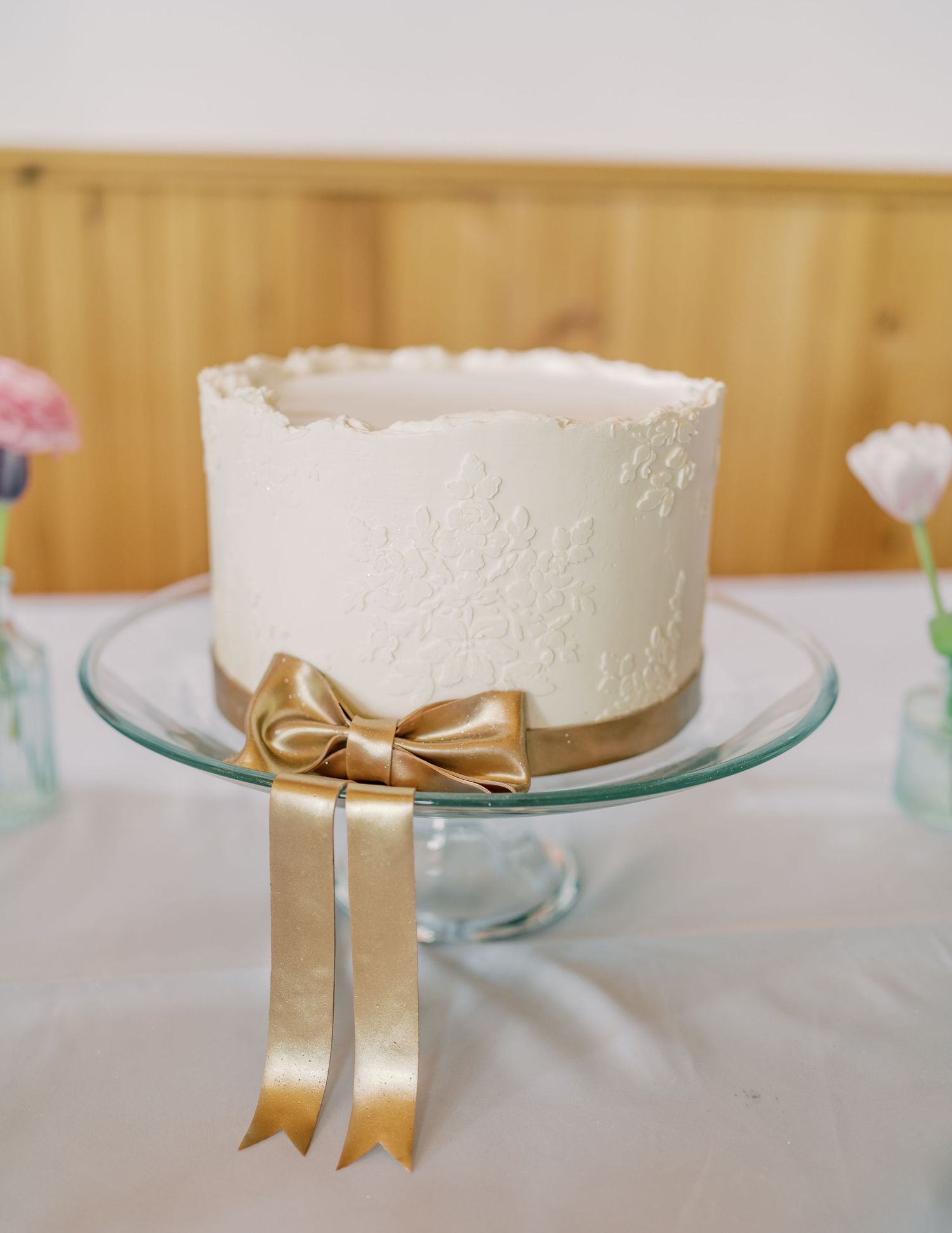 A white wedding cake decorated with delicate lace patterns and a gold bow ribbon sits on a glass cake stand.