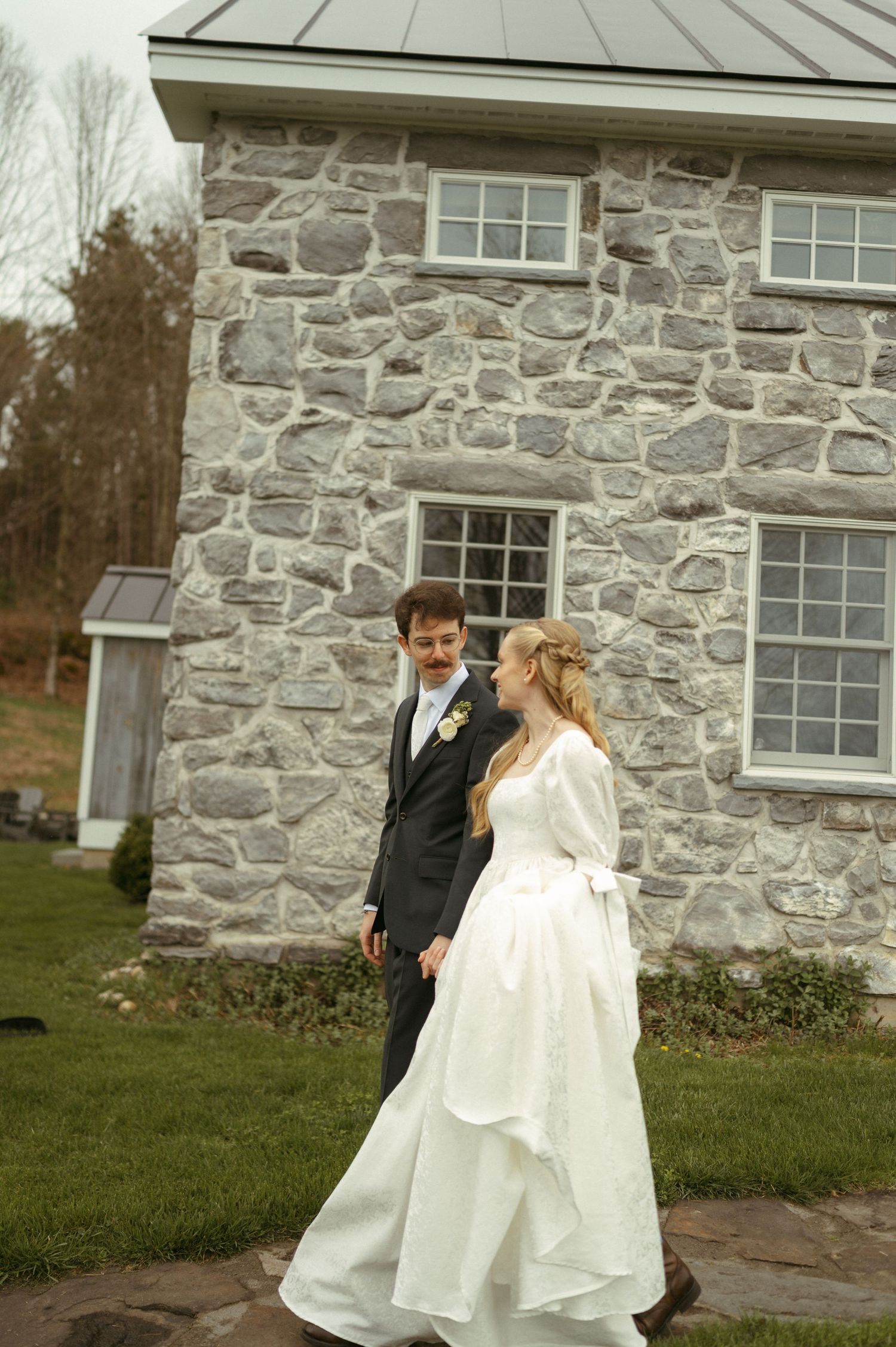 A couple in wedding attire share a playful moment outside a stone building.
