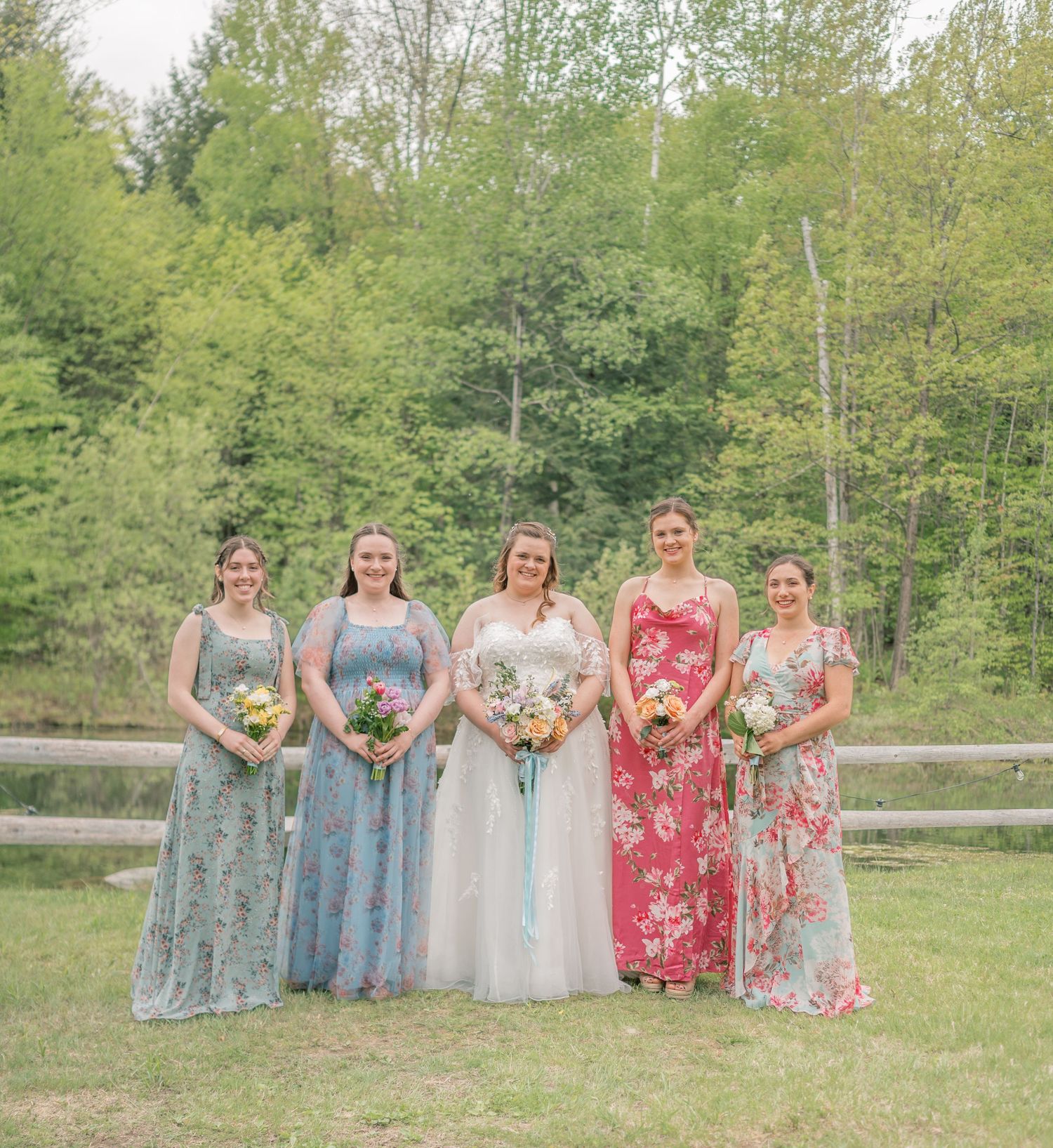 A group in formal floral dresses stands together on grass with a wooden fence and green trees in the background.