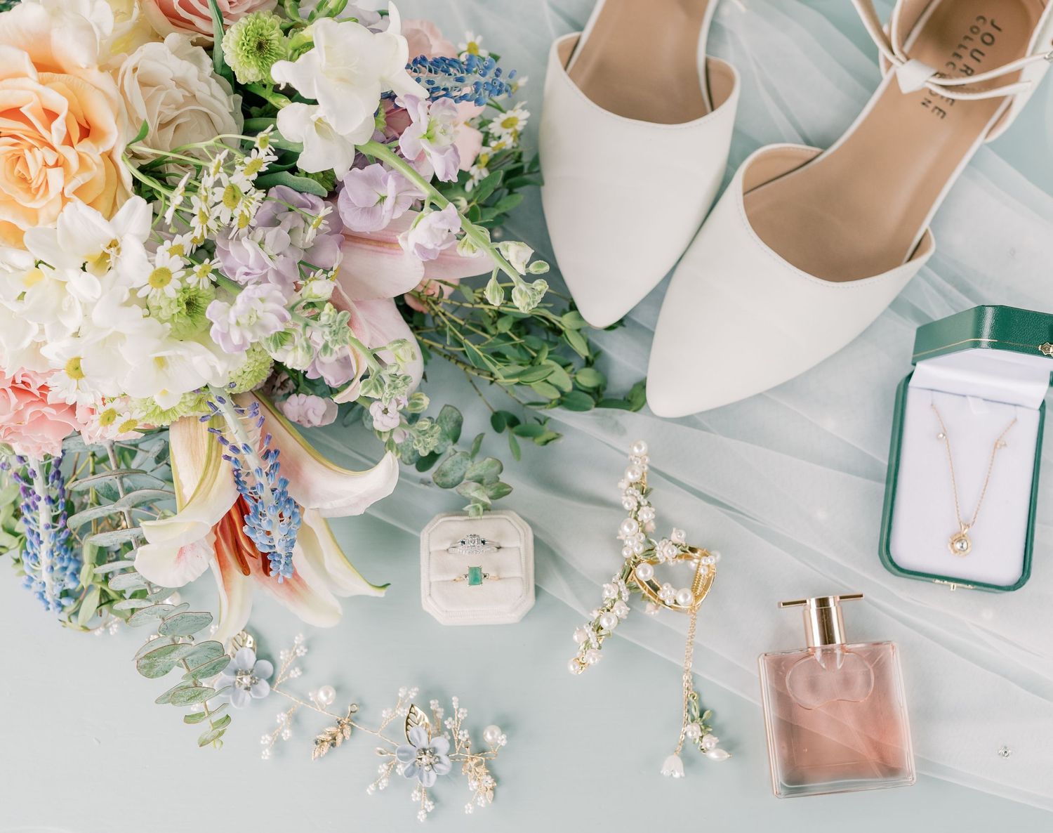 Wedding accessories including white heels, delicate floral arrangements, and perfume bottle arranged on light blue surface.
