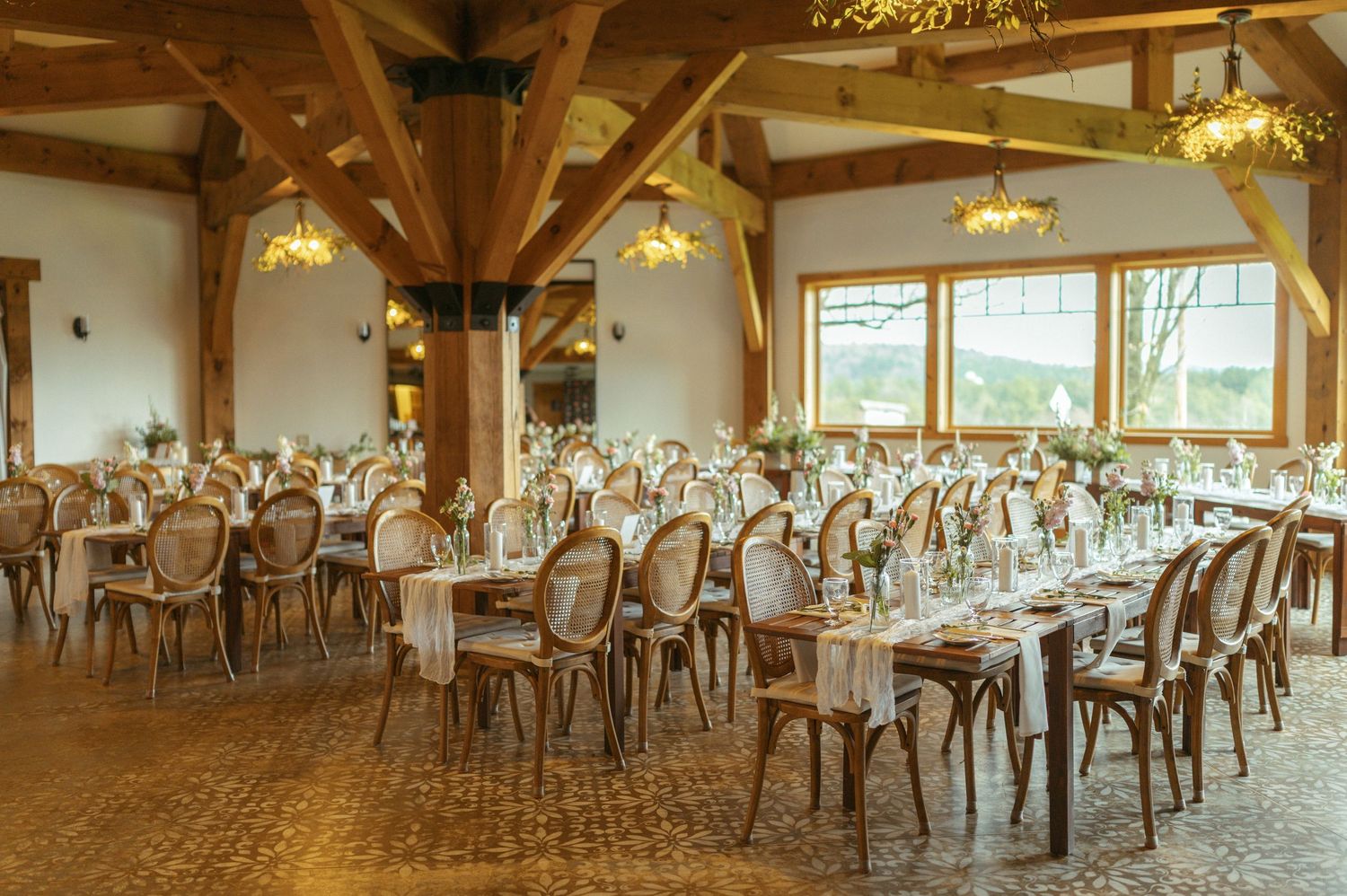 Large rustic event venue with exposed wooden beams, chandeliers and elegant table settings for a wedding reception.