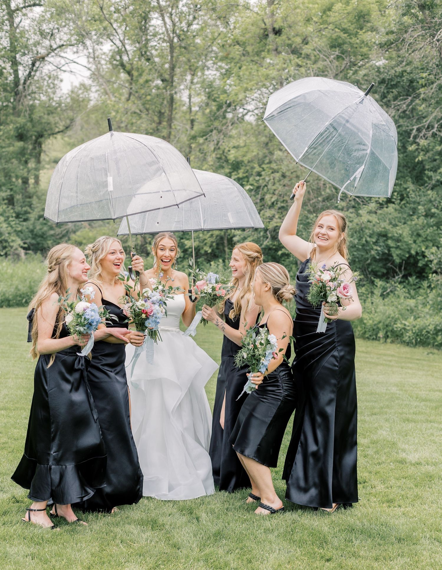 Bridal party in navy dresses and white gown pose playfully with clear umbrellas on a green lawn.