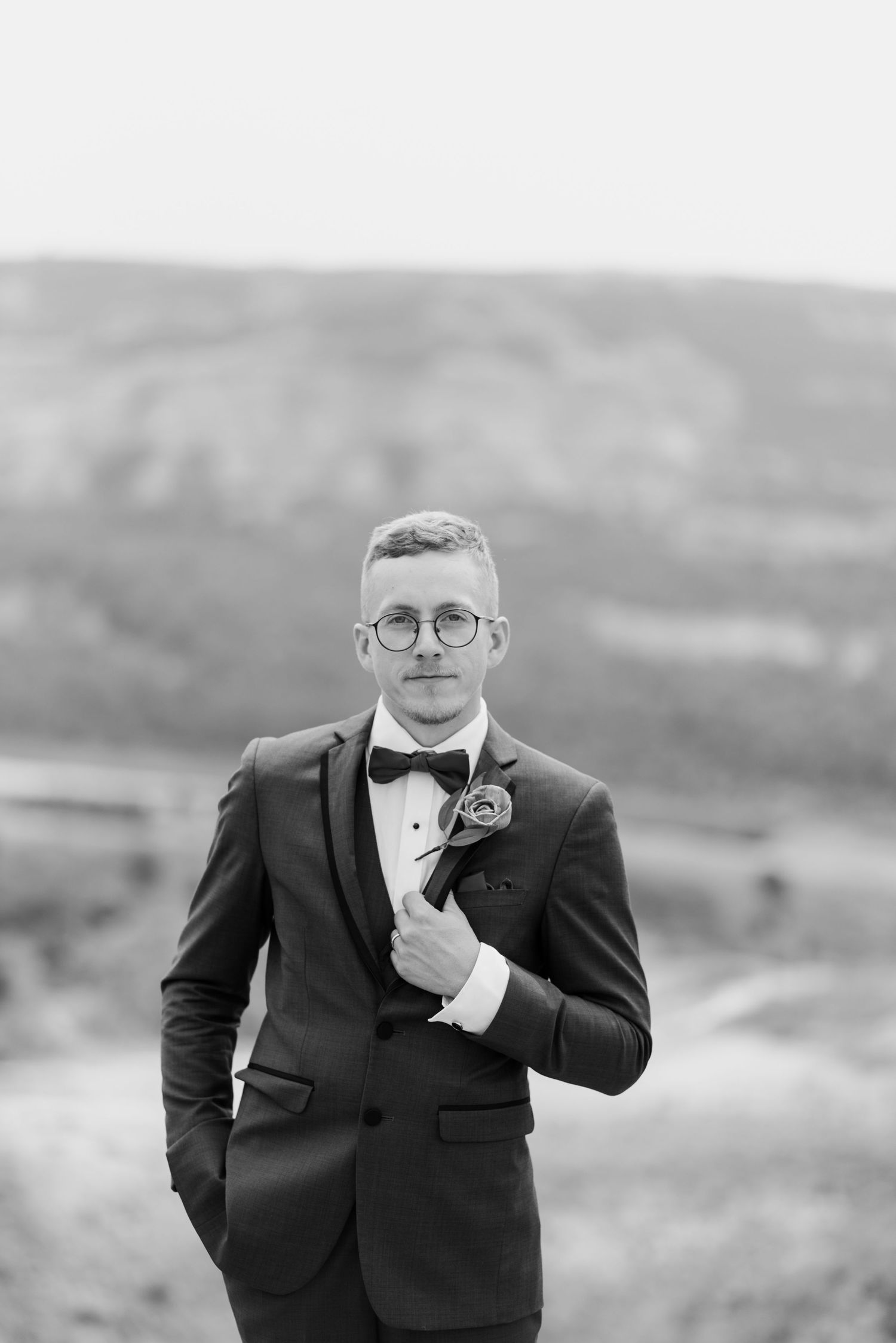 A groom in a black tuxedo and bowtie poses emotionally during an outdoor wedding photoshoot.