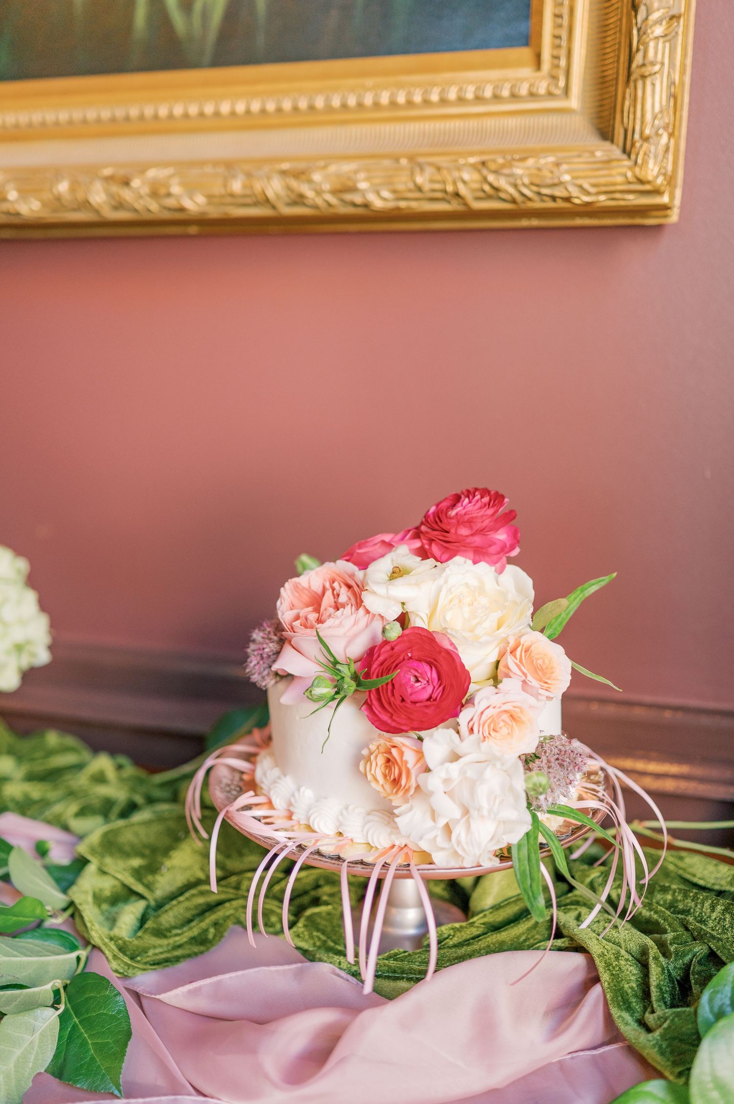 Pink and coral roses adorn a delicate one-tiered cake, part of a traditional cake pull with satin ribbons against a pink wall background.