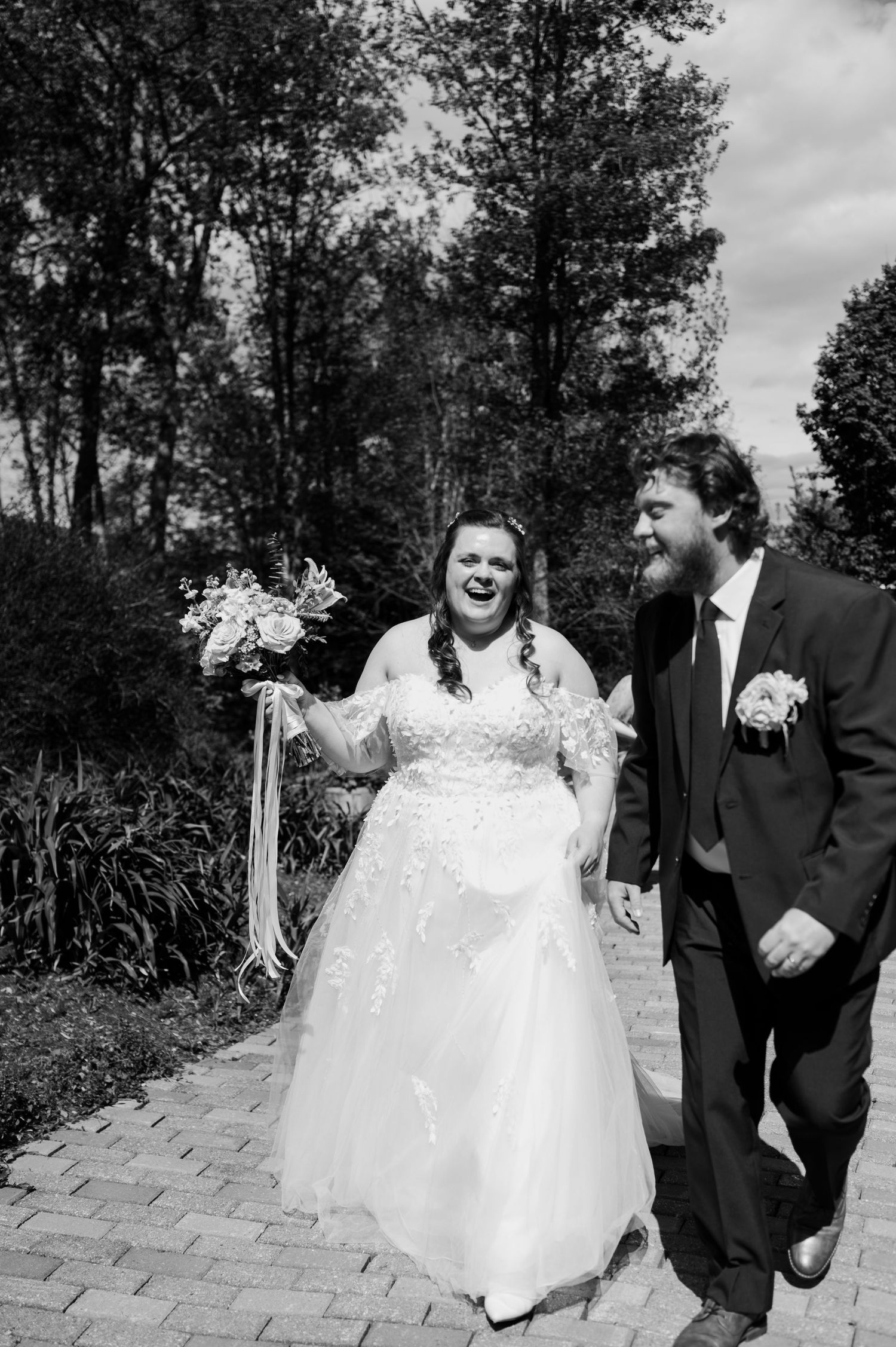 A joyful wedding couple walks together on a stone path, with trees in the background captured in black and white.