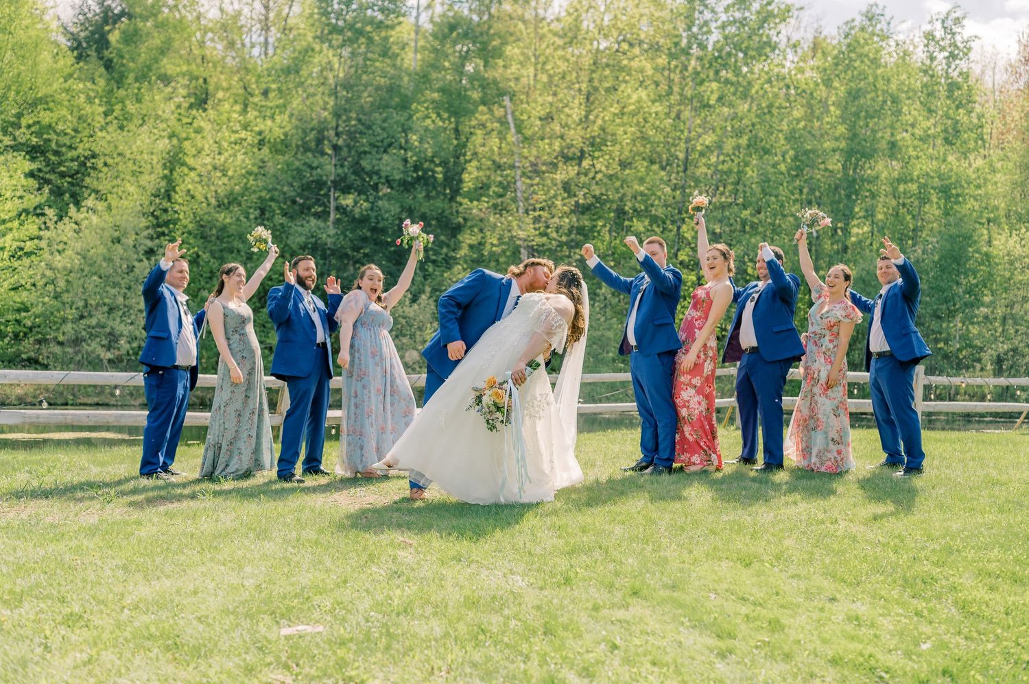 Wedding party stands in line on green grass with trees in background during outdoor summer ceremony.