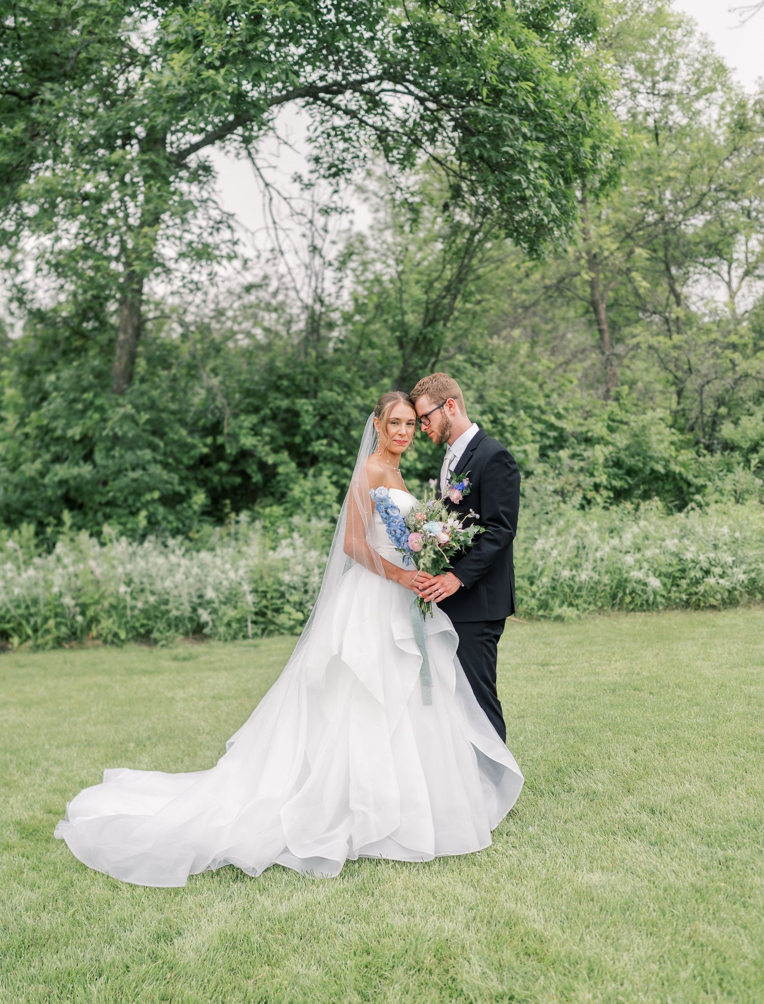A couple shares a romantic moment on their wedding day in a lush garden setting with a flowing white gown and blue and white floral bouquet.
