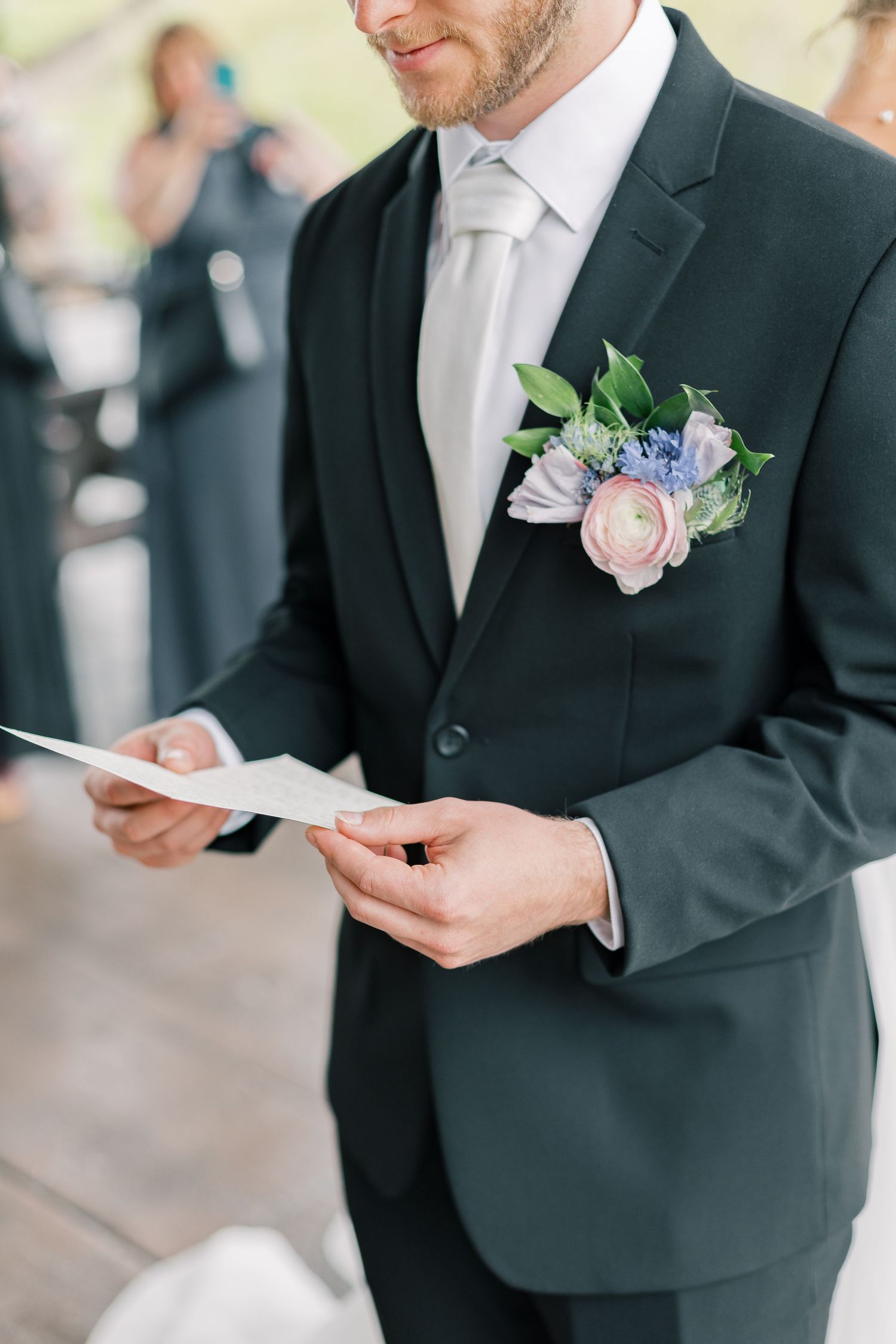 Groomsmen in black suits wearing delicate floral boutonnieres read from papers during a wedding ceremony.