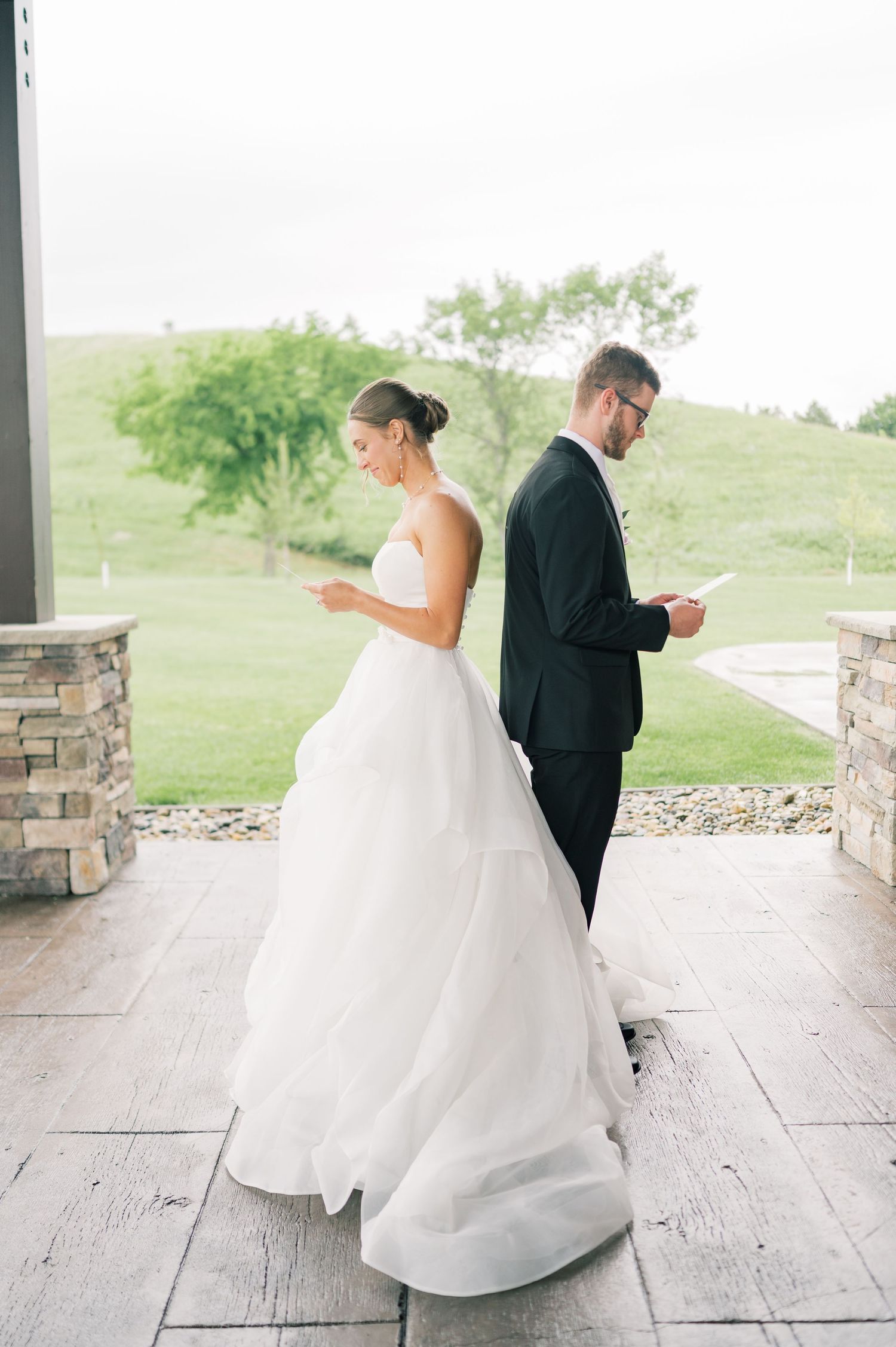 A bride and groom stand back-to-back on a stone patio while checking their phones in a candid wedding moment.