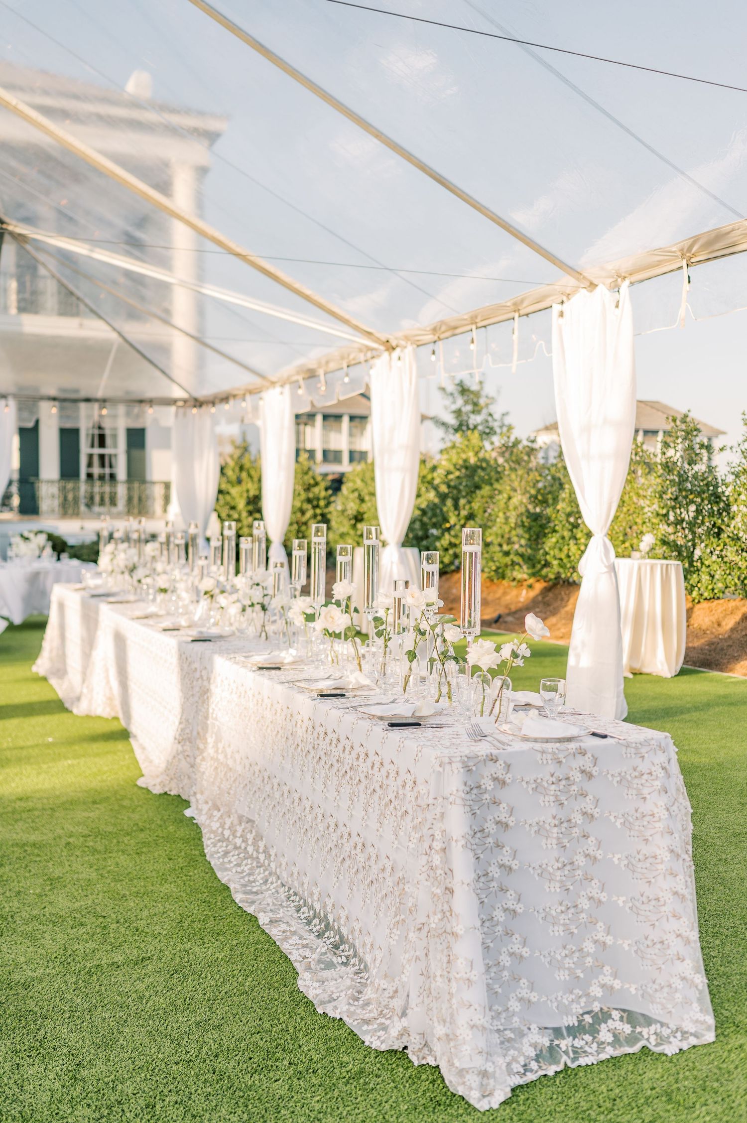 Elegant outdoor wedding reception table with white lace tablecloth and sheer curtains under a clear tent on manicured lawn.