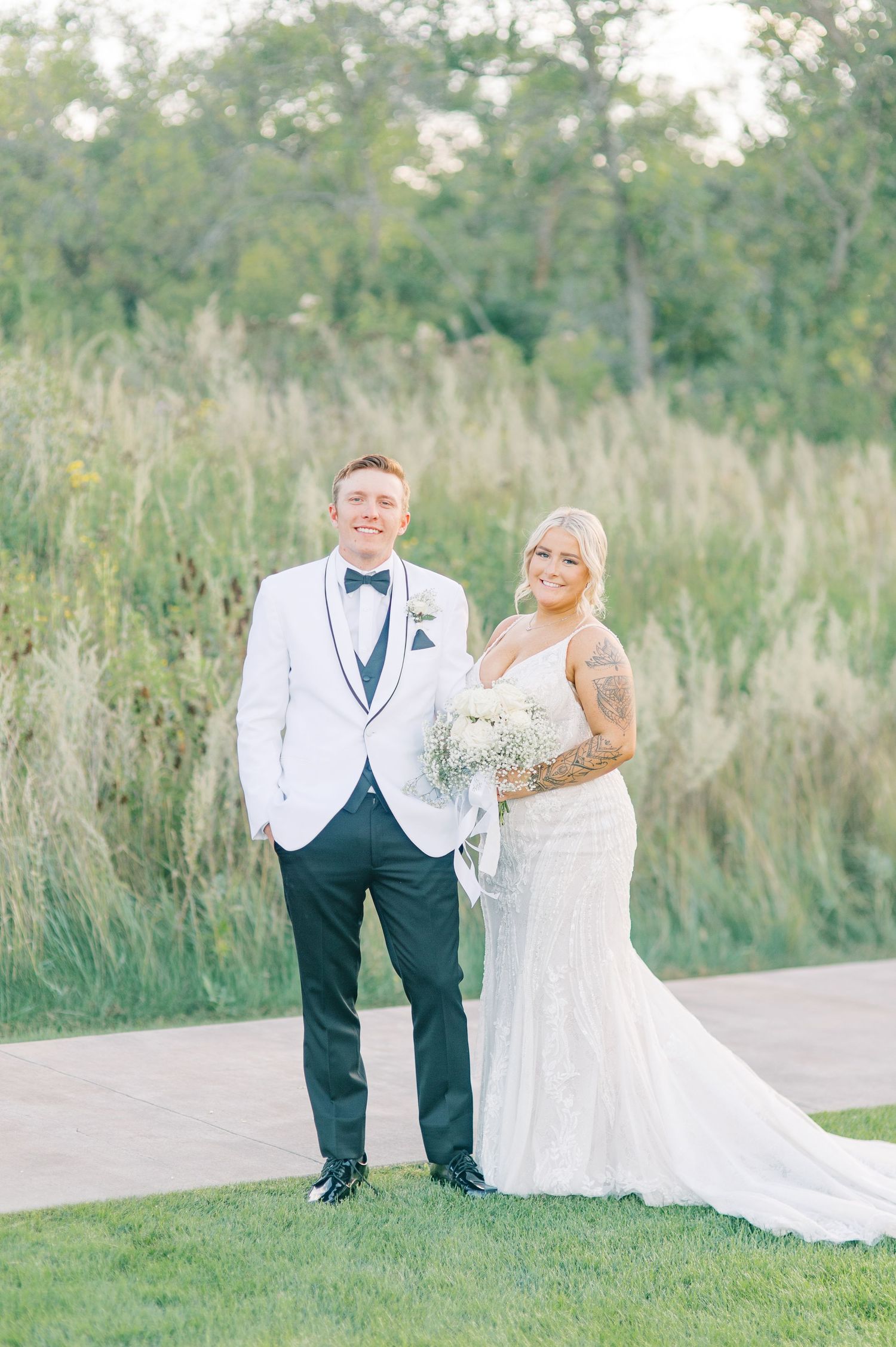 A wedding couple poses together outdoors in formal attire with tall grass and trees in the background.