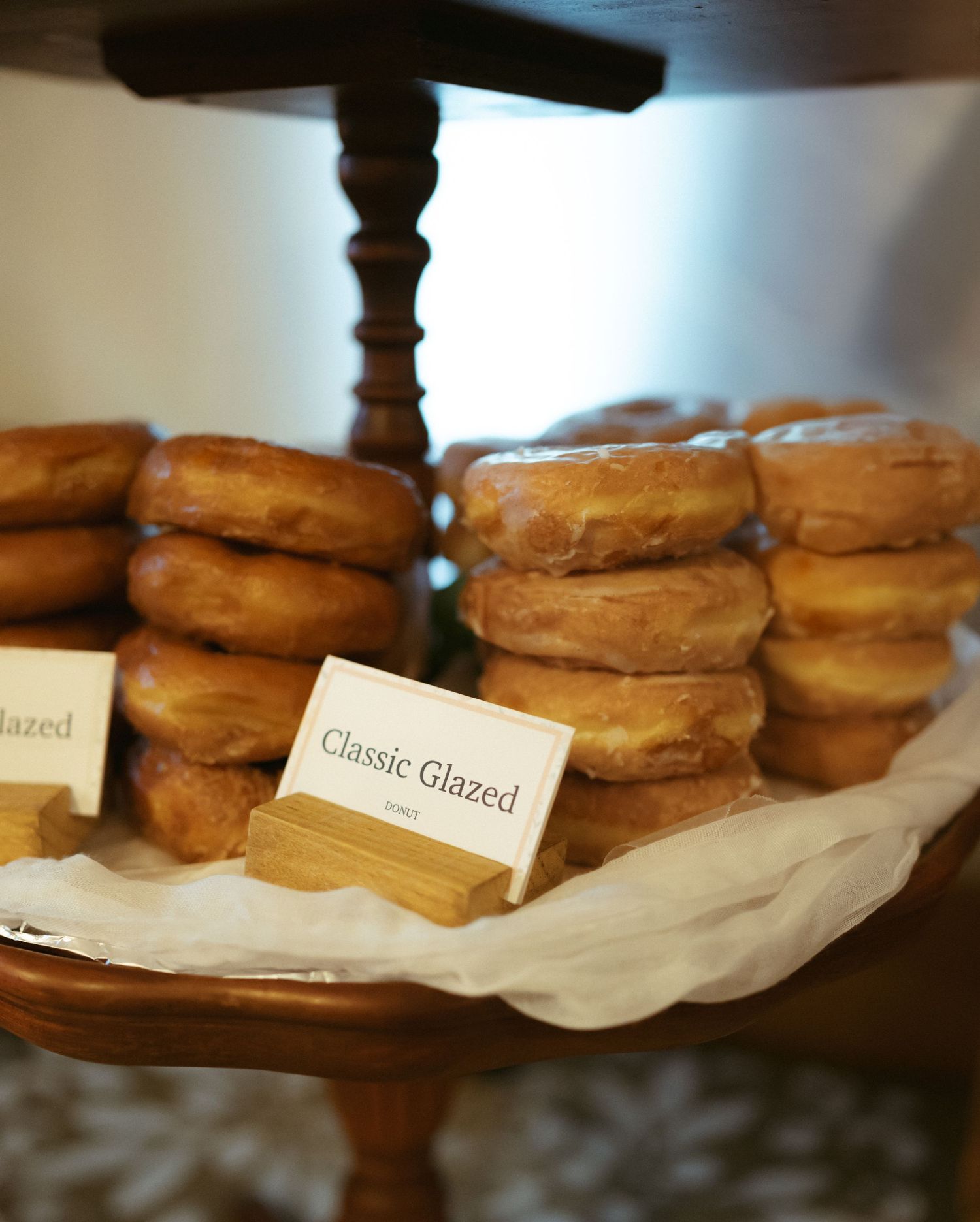 Stacks of glazed donuts displayed on a tiered wooden stand with a small label reading 'Classic Glazed'.
