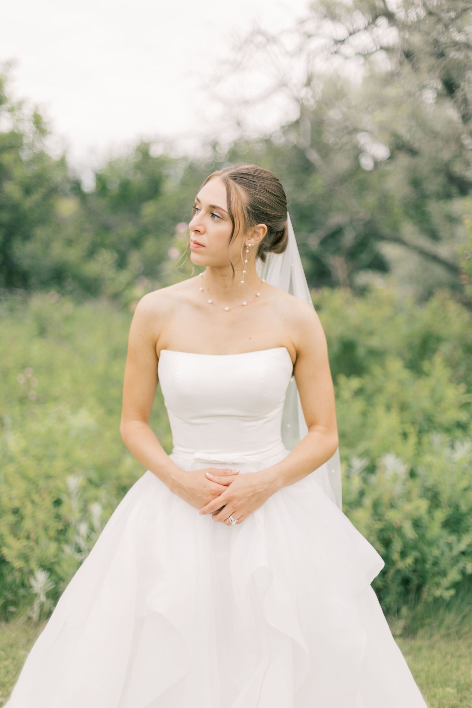 A bride in a strapless white ballgown and veil poses outdoors in a natural garden setting with soft lighting.