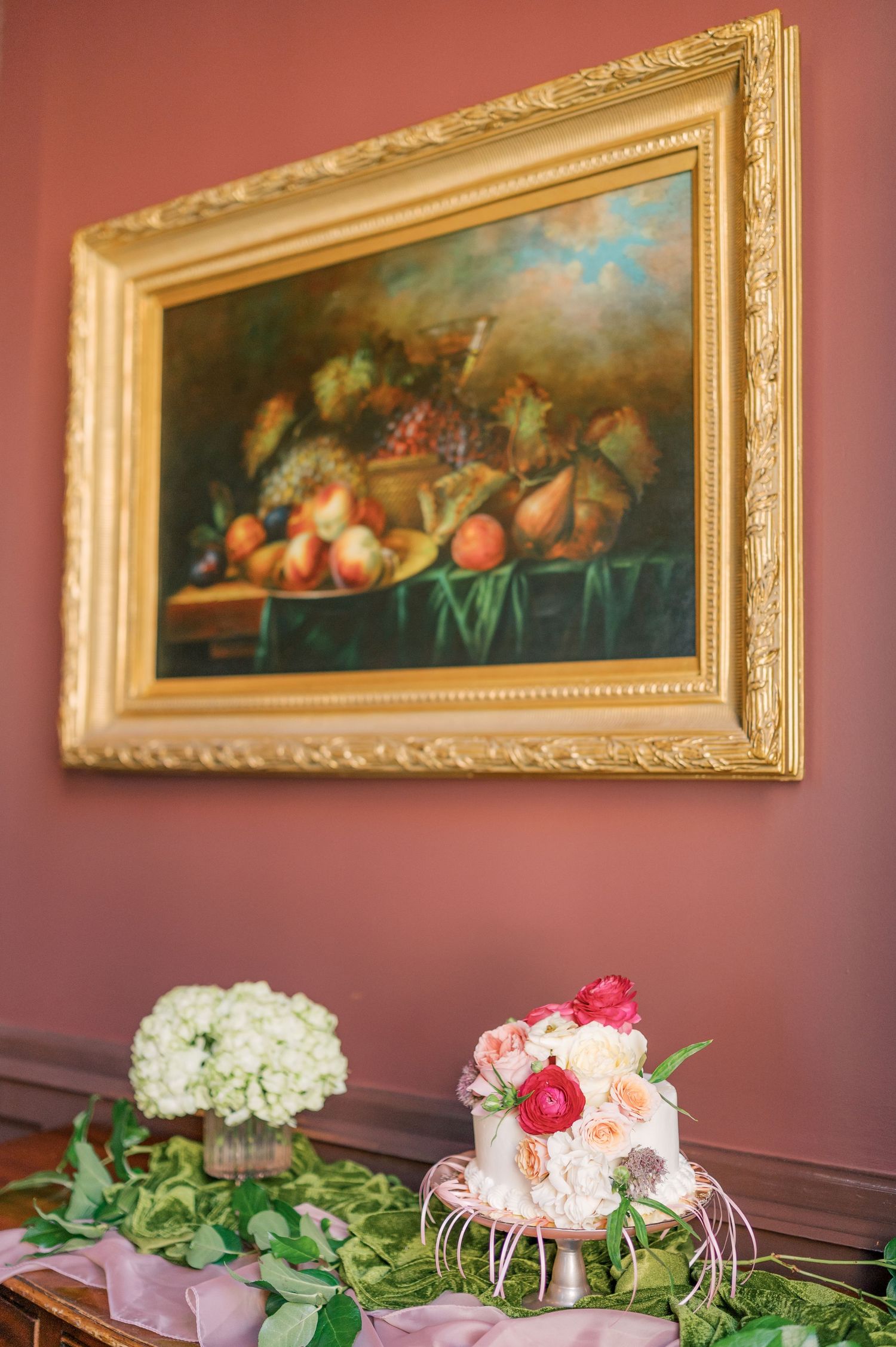 Elegant still life painting in gold frame above table with pink and white floral arrangements and greenery.