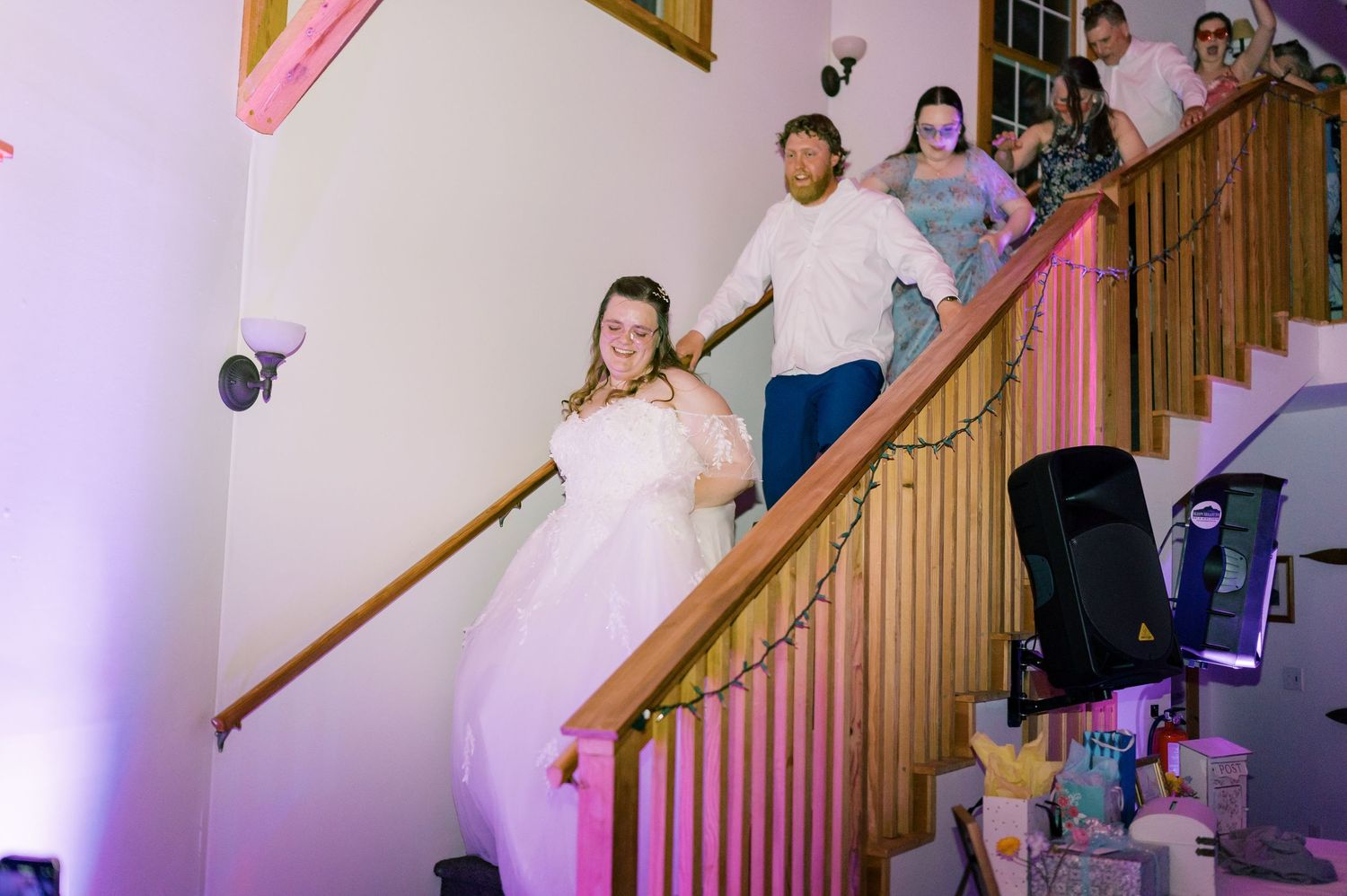 A group of people dance and celebrate while descending a wooden staircase with pink lighting.