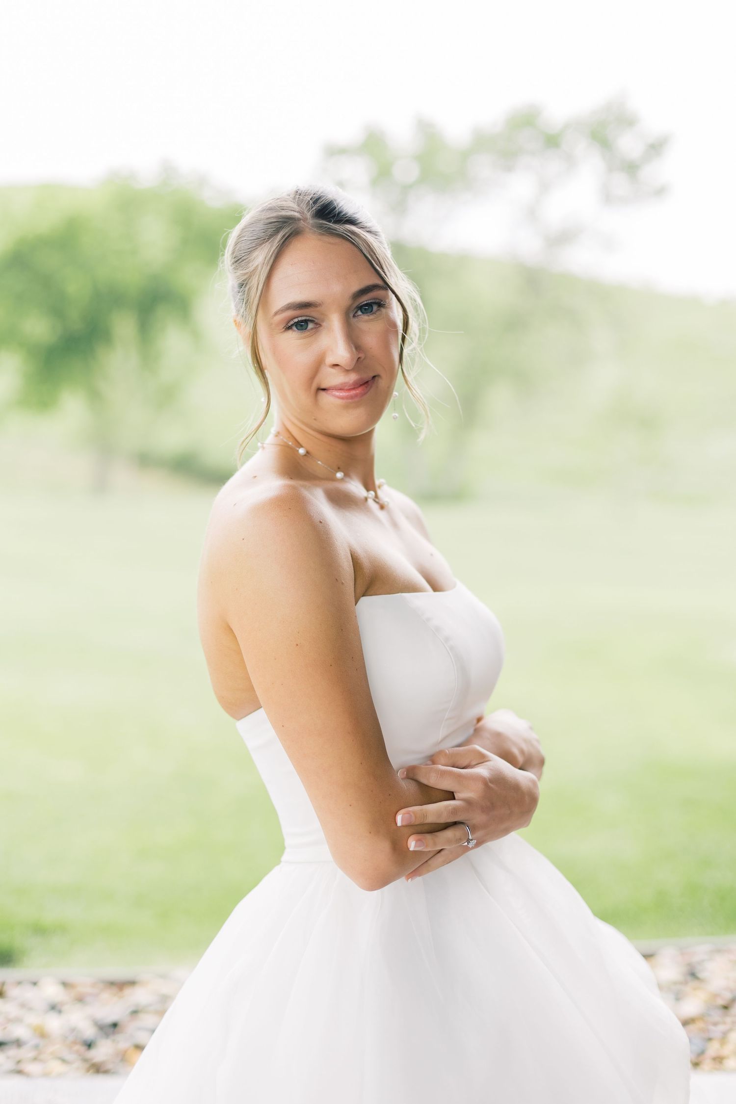 A bride in a white ballgown poses outdoors in a dreamy natural setting with soft lighting and greenery in the background.