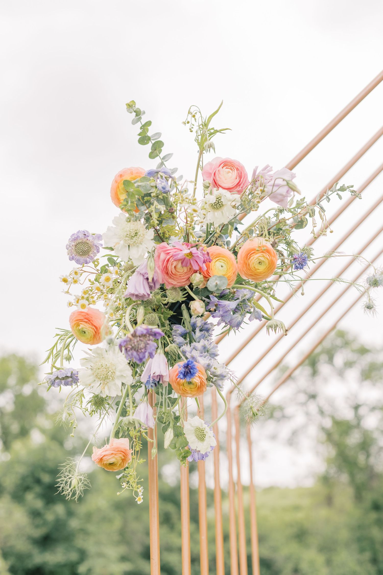 Delicate floral arrangement with peach ranunculus and purple blooms adorns a wooden arch against a soft outdoor backdrop.