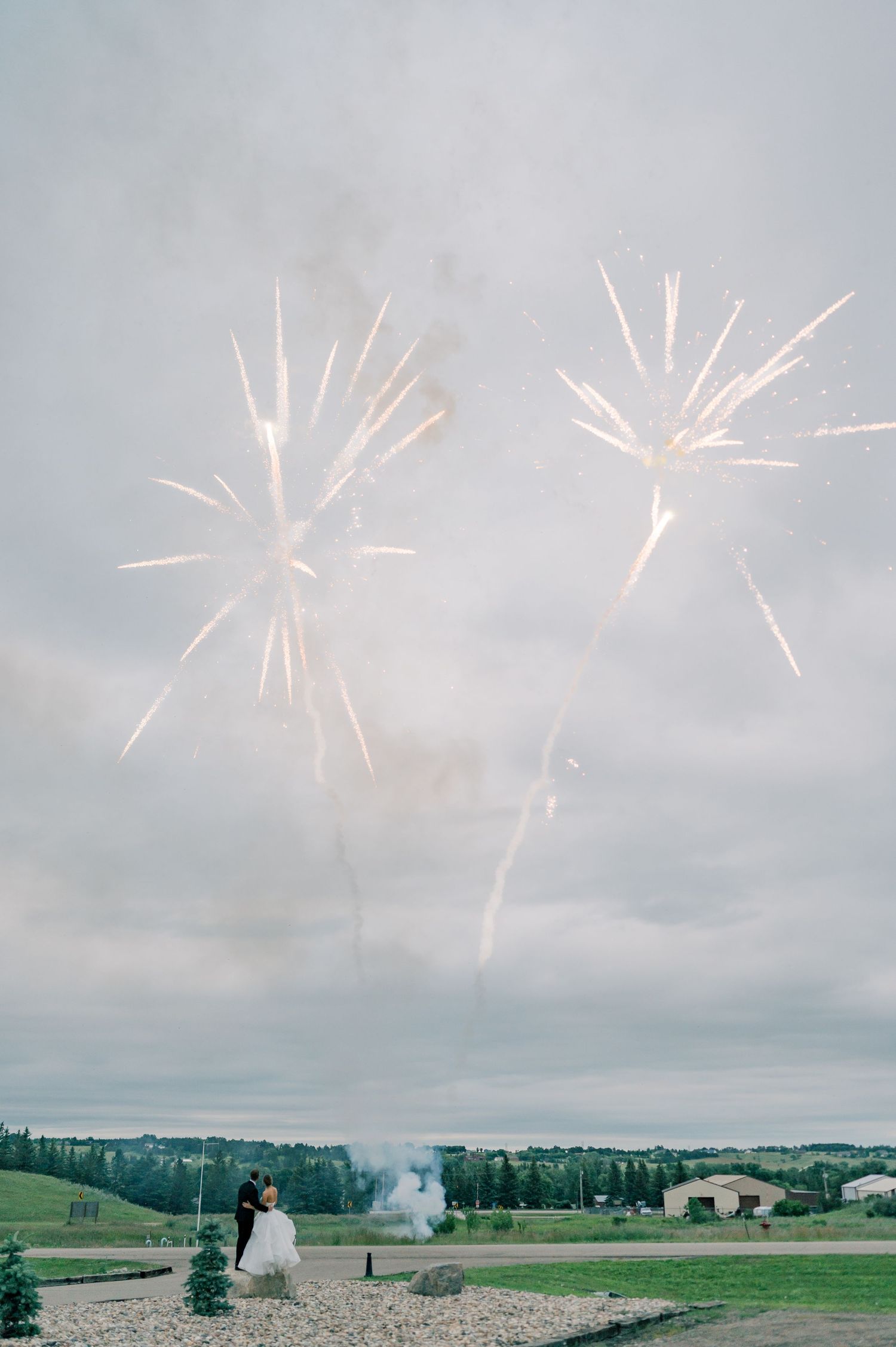 Pink fireworks explode in a cloudy sky over a green rural landscape with scattered buildings and trees.