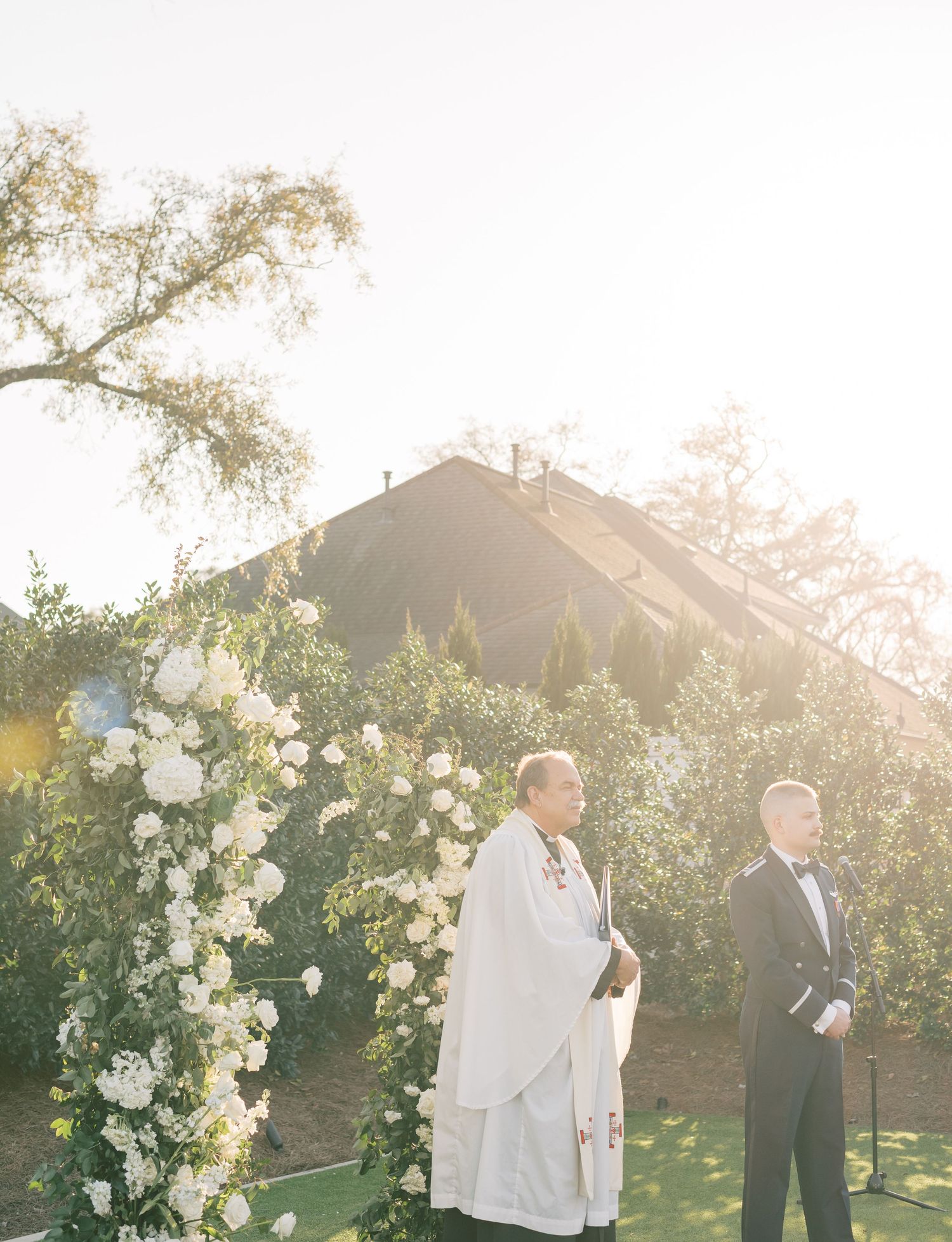 Groom and pastor with white floral arrangements during golden hour sunset.