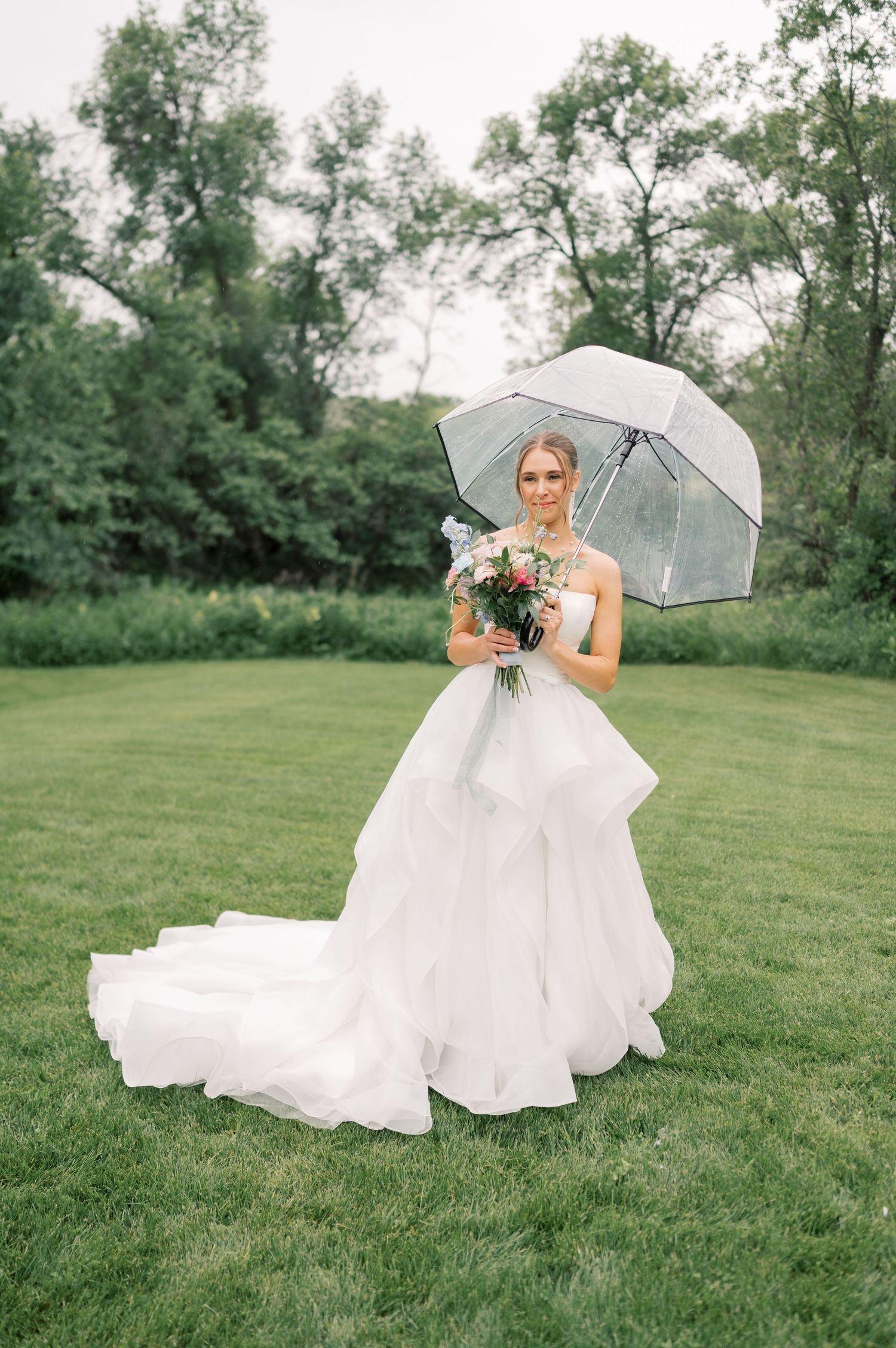 A bride in a flowing white dress sits gracefully on lush green grass while holding a clear umbrella on a cloudy day.