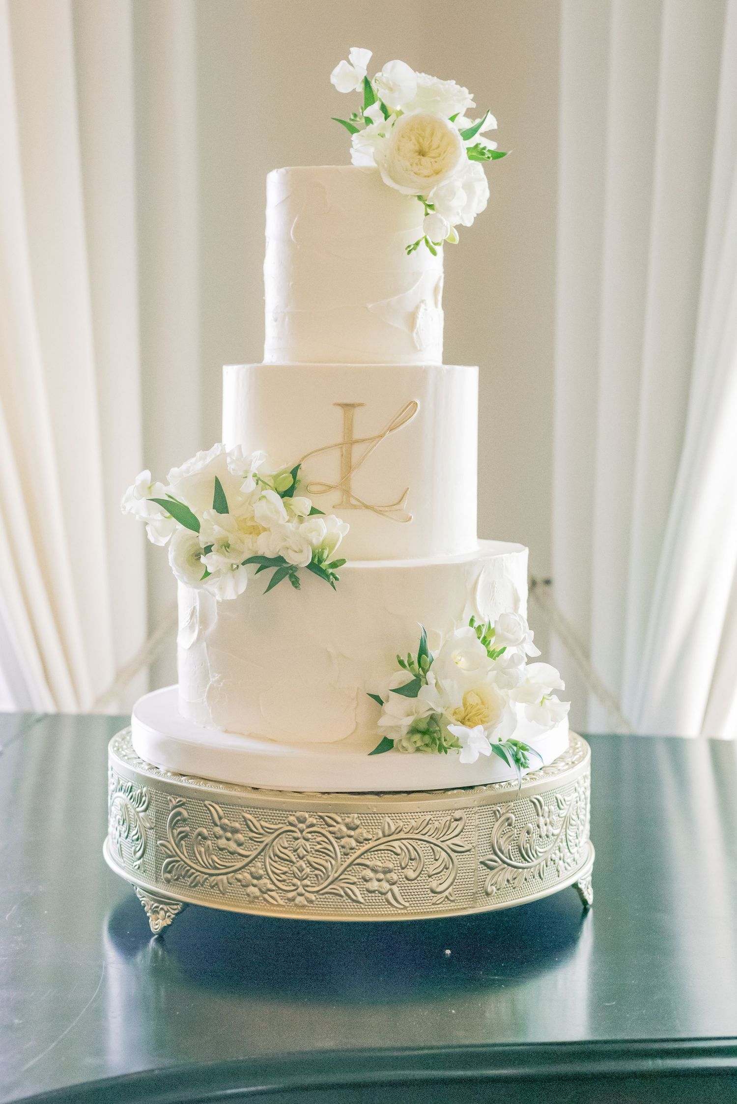 White tiered wedding cake with delicate floral decorations and ornate silver base displayed against white curtain backdrop.