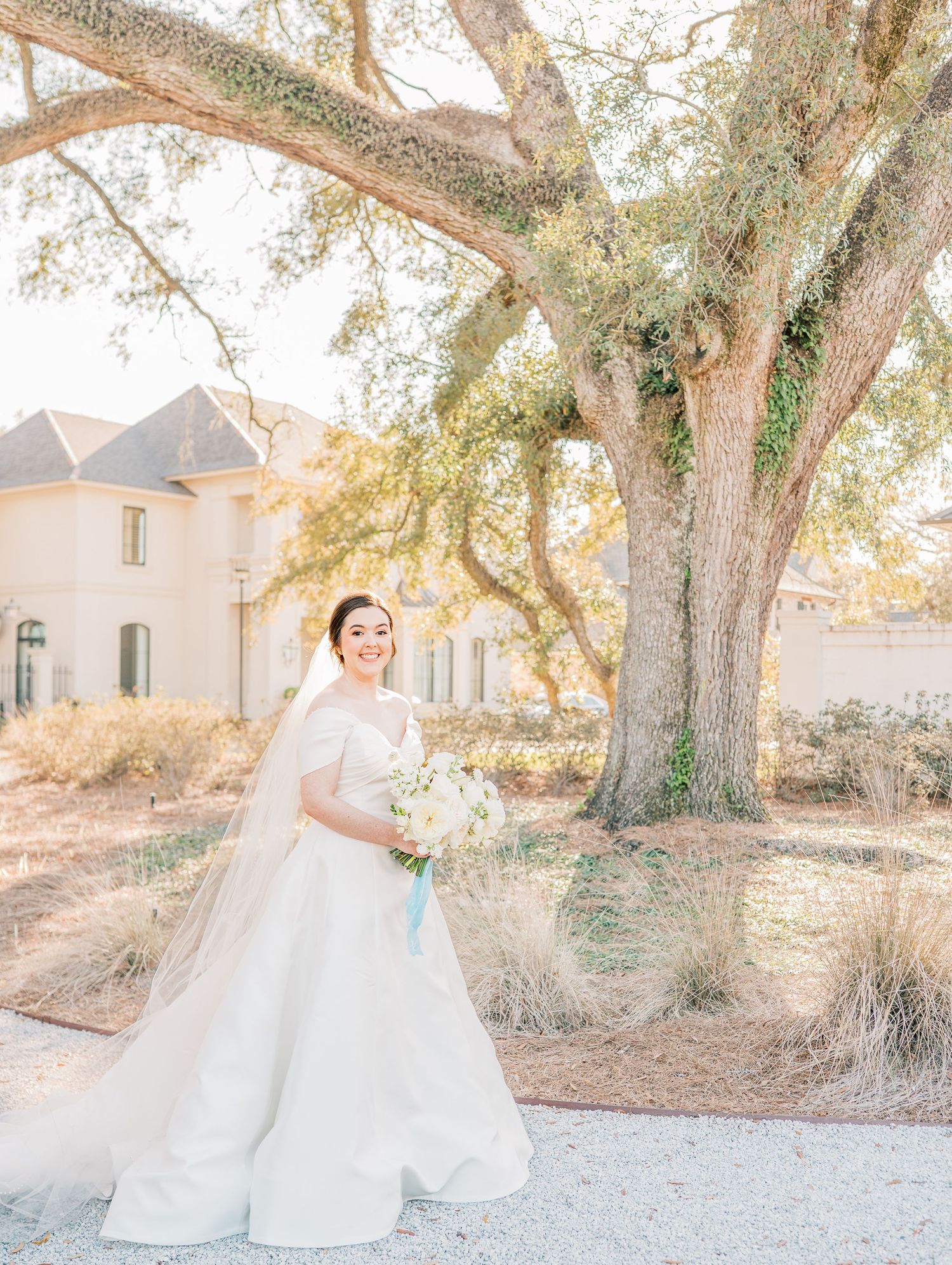 A bride in a flowing white gown poses elegantly under majestic oak trees at an outdoor wedding venue.