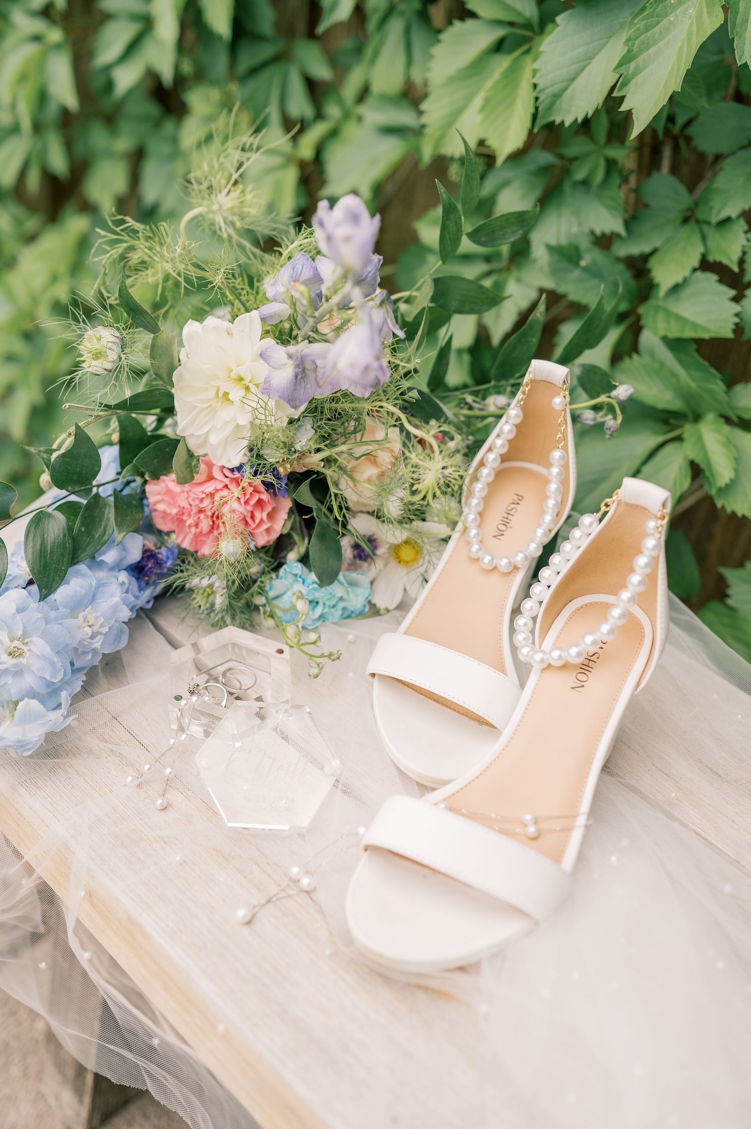 Delicate floral arrangements surround elegant beige sandals on a stone surface with greenery in the background.