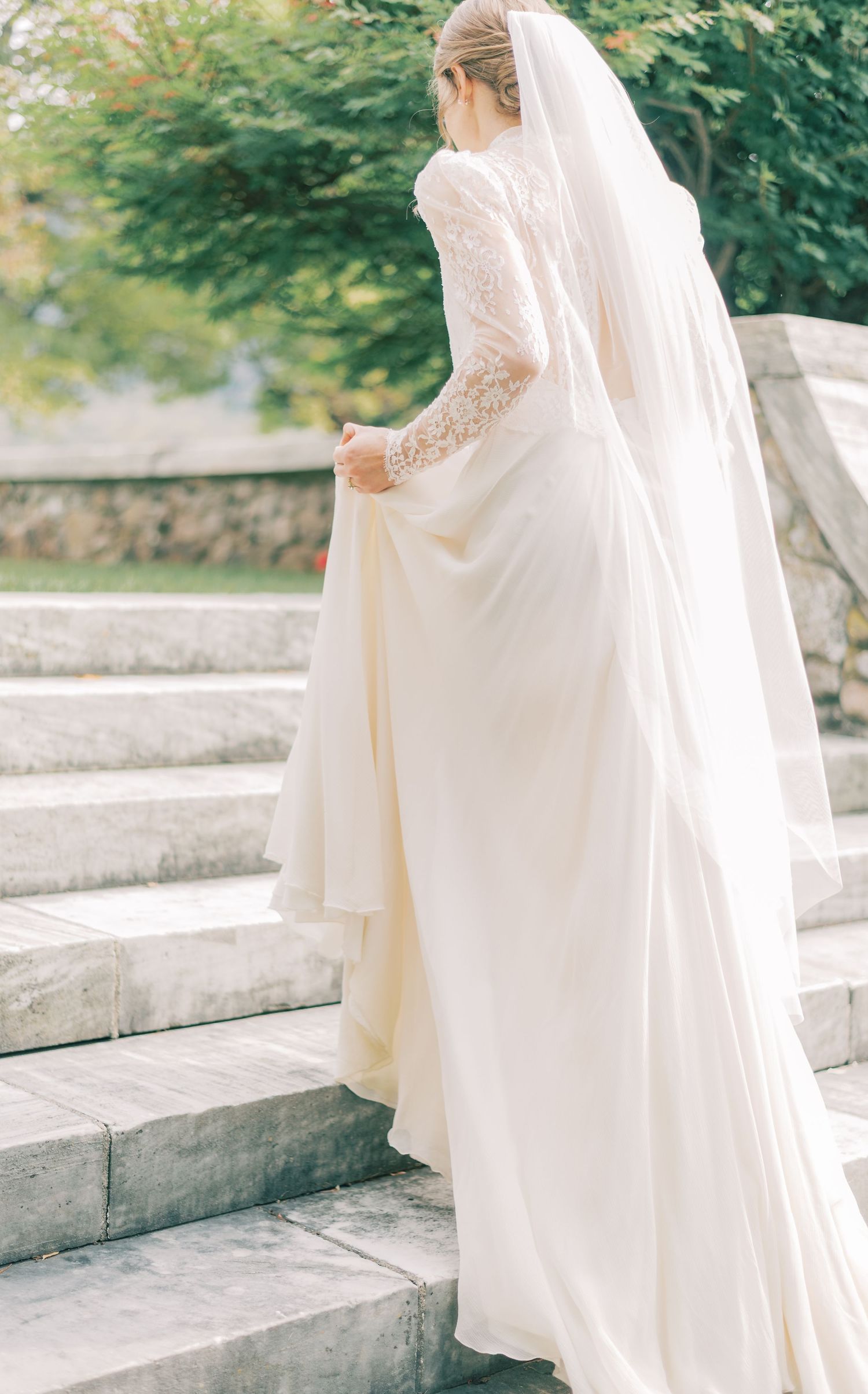 A bride in a flowing white wedding dress and veil walks up stone steps in a garden setting.