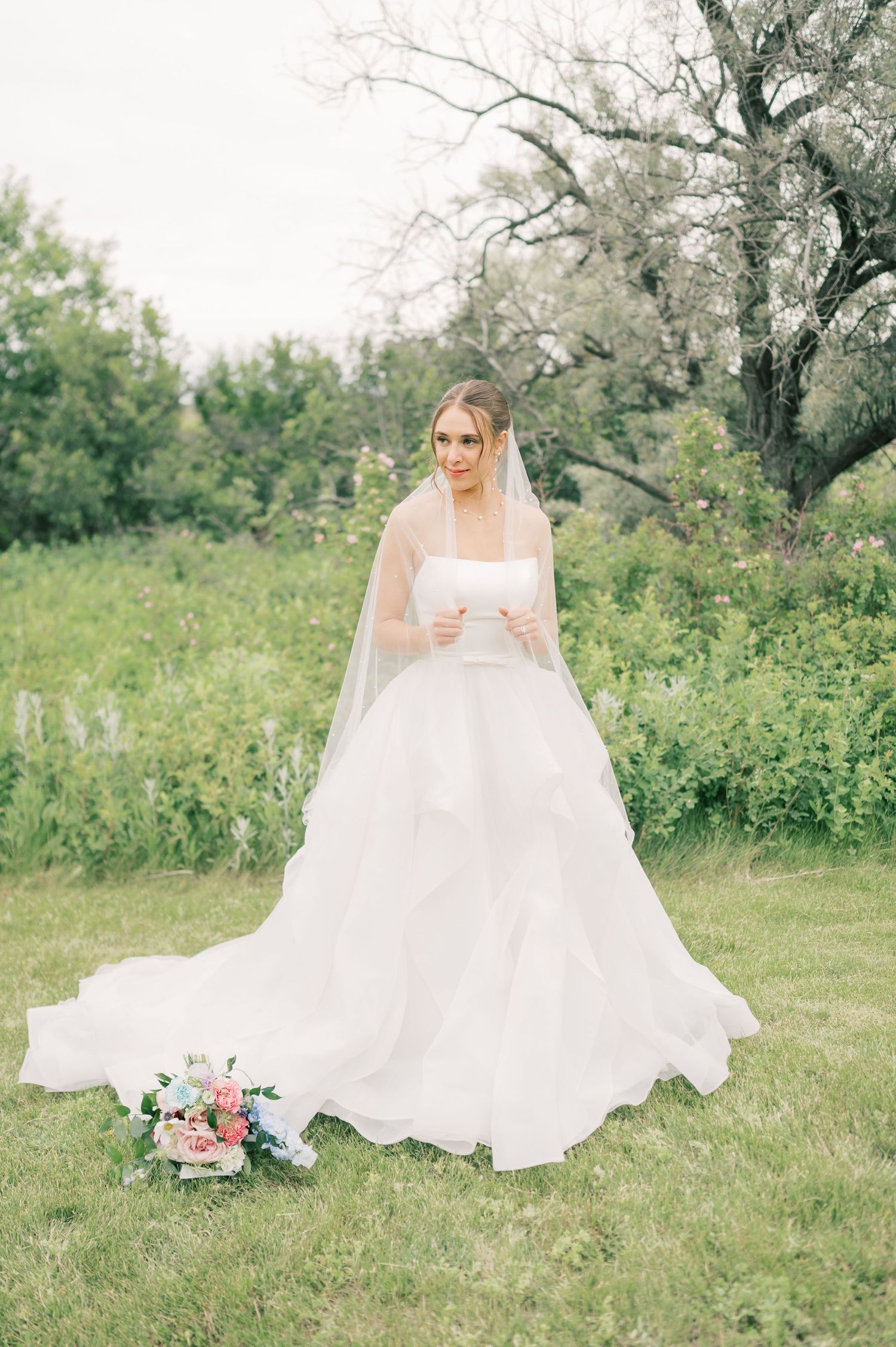 A bride in a flowing white wedding gown sits gracefully in a spring meadow surrounded by blooming trees and flowers.