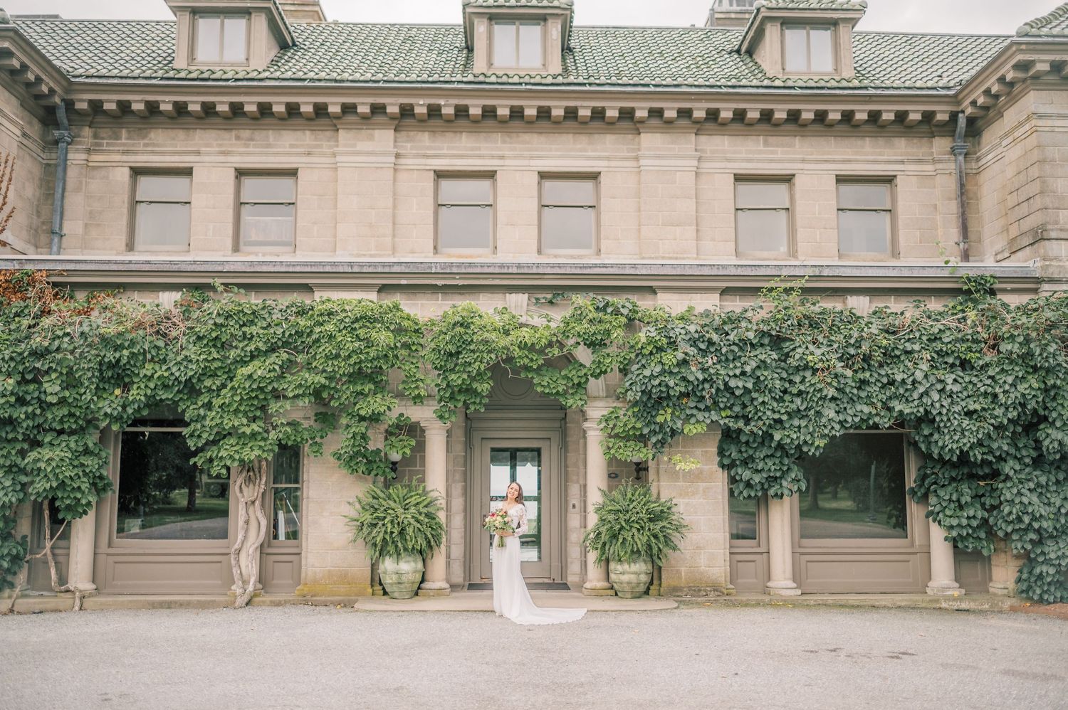 Elegant bride in white dress stands between ivy-covered columns at a grand historic mansion entrance.