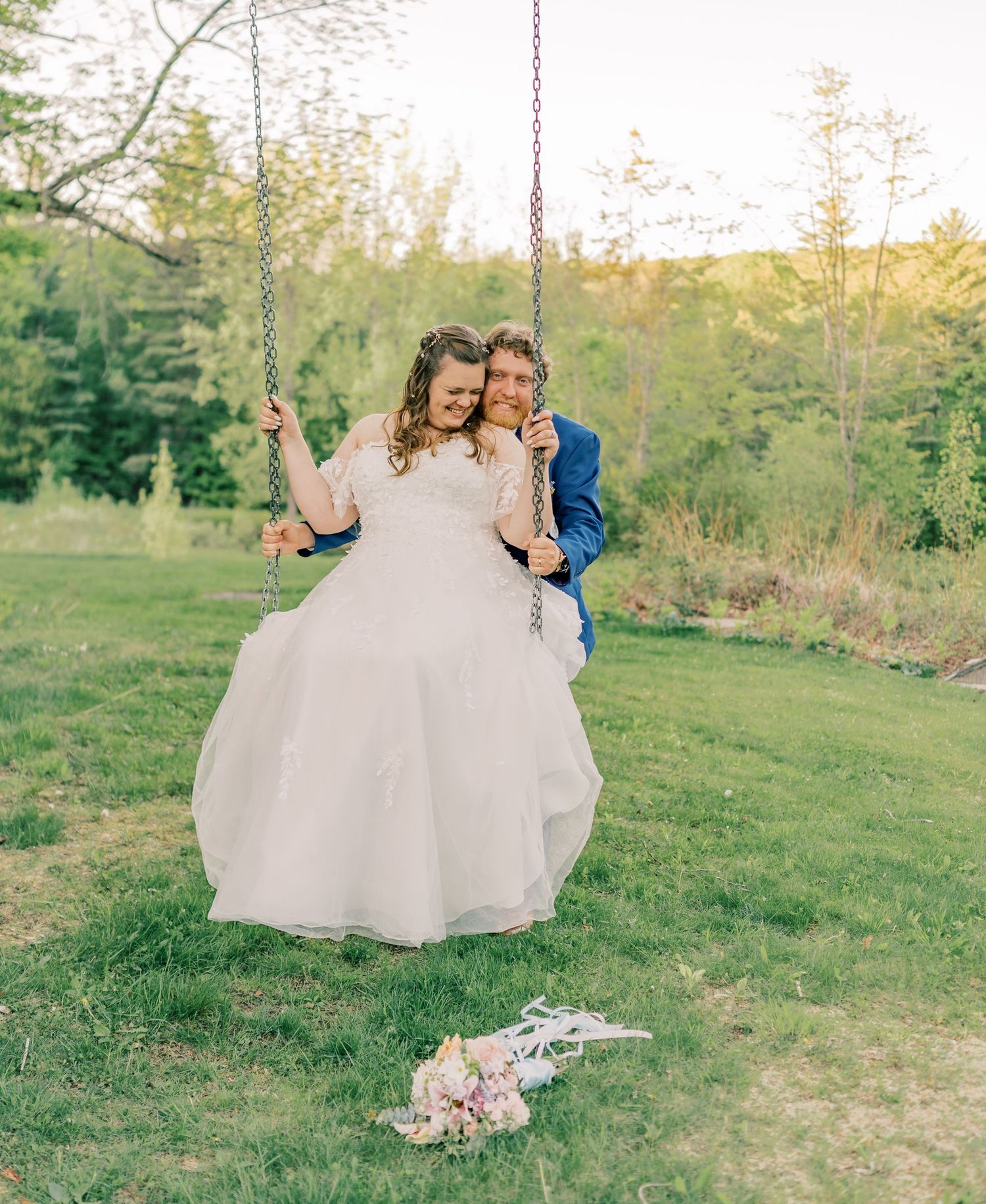A couple in wedding attire shares a romantic moment in a sunlit meadow with trees in the background.