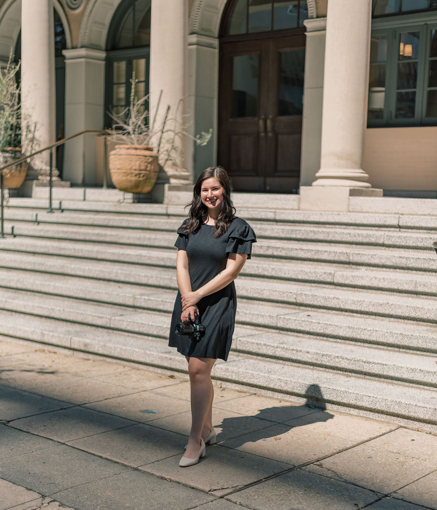 A woman in a black dress and heelss poses on stone steps in front of a building with classical columns on a sunny day.
