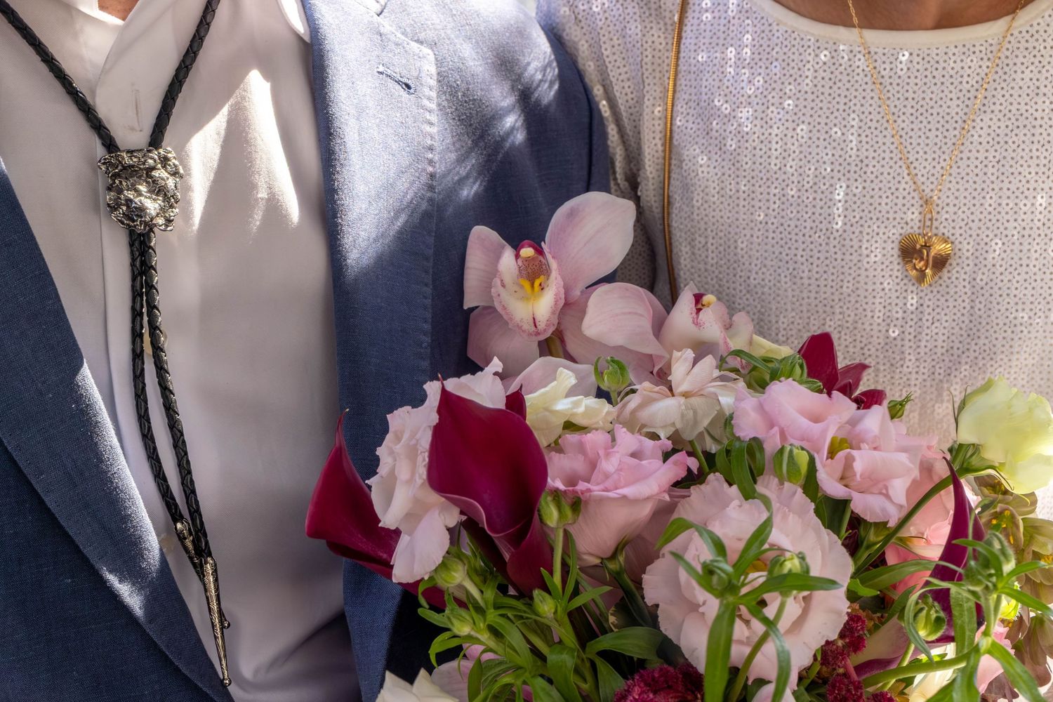 Close-up of a couple holding a bouquet of pink and red flowers, with a bolo tie and gold necklace visible.
