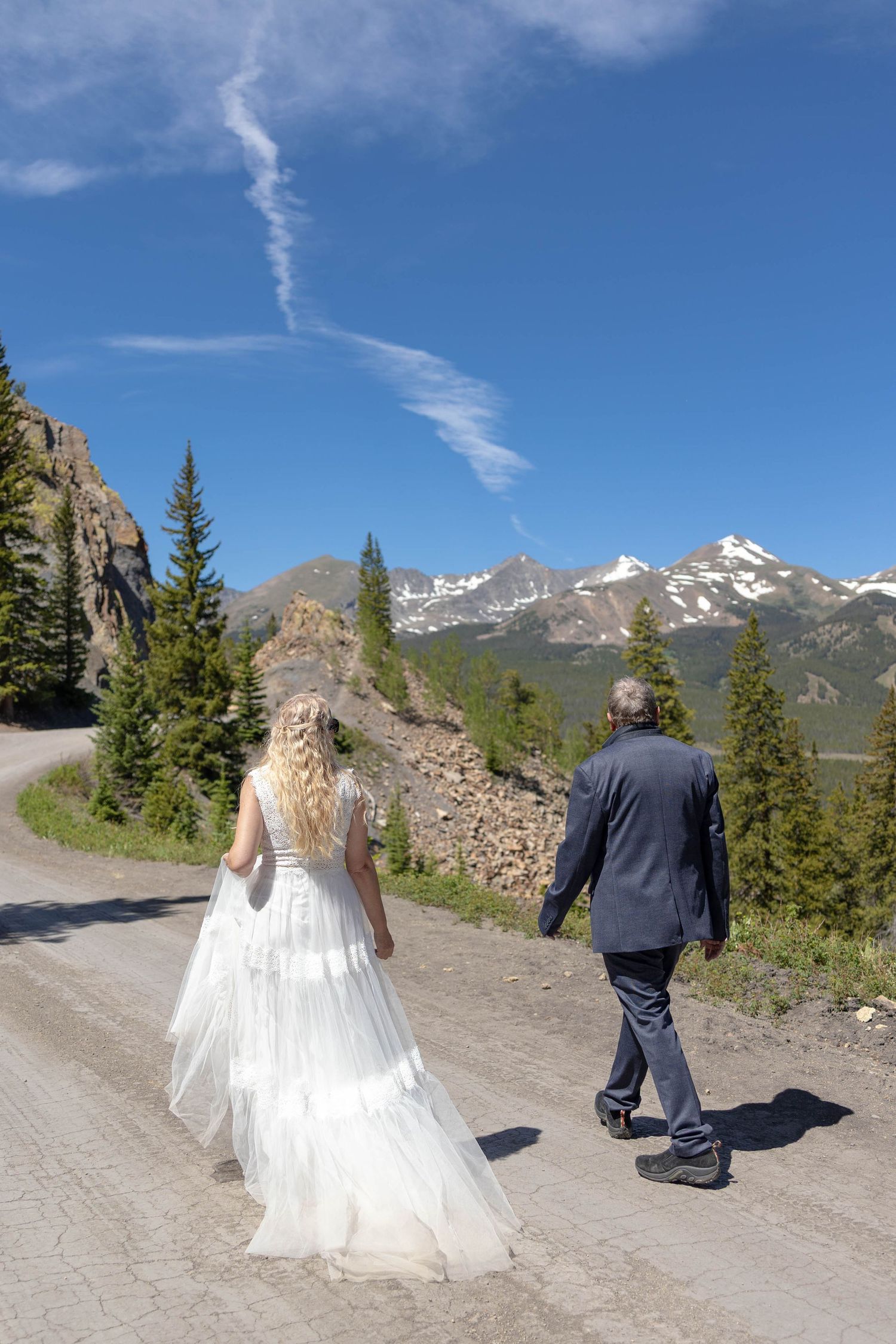 Bride and groom walking hand in hand on a mountain road with snow-capped peaks and pine trees under a blue sky.